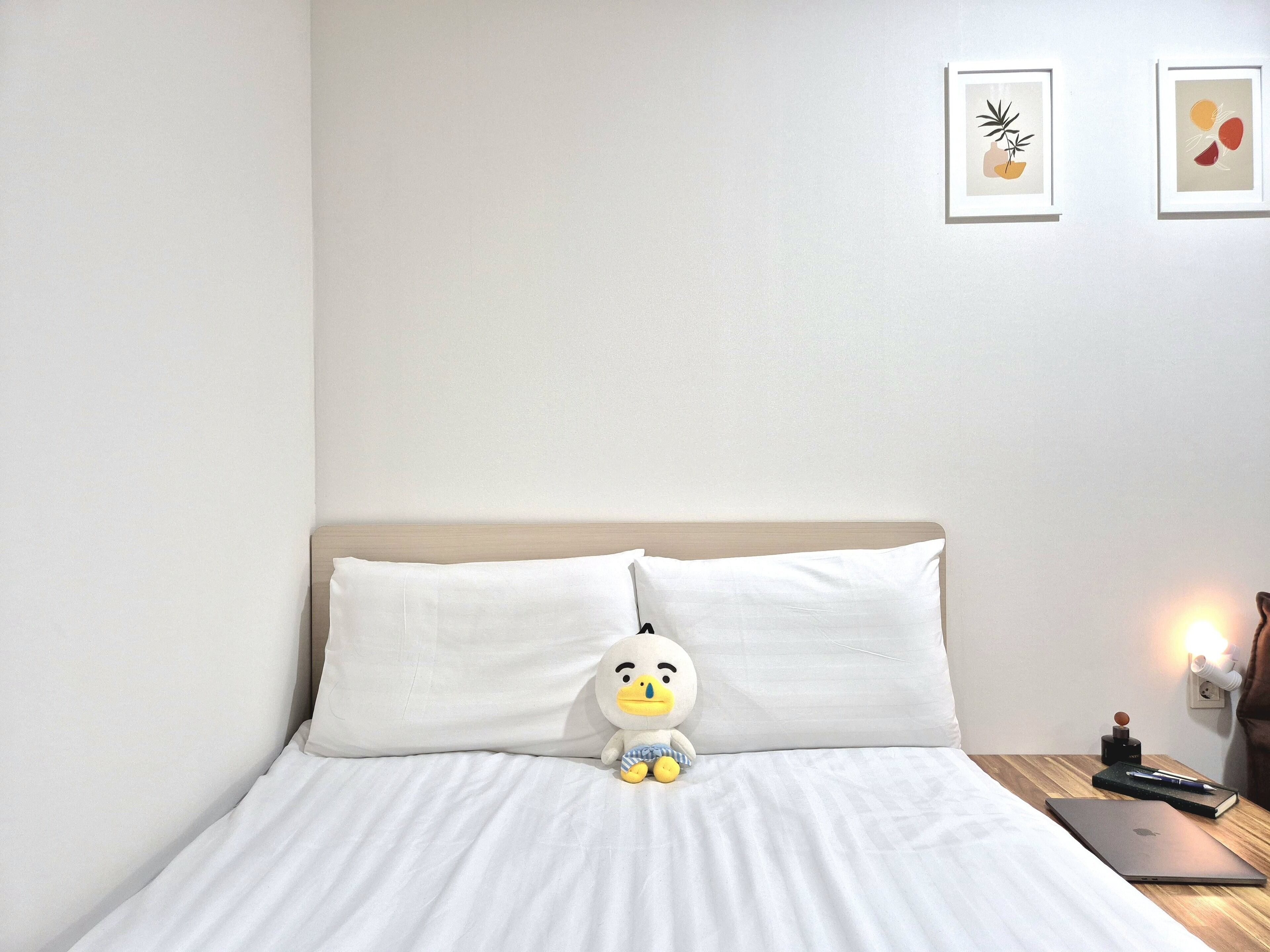 A neatly made bed with white linens, a plush toy duck, a laptop, and a nightstand with a lamp and a book.