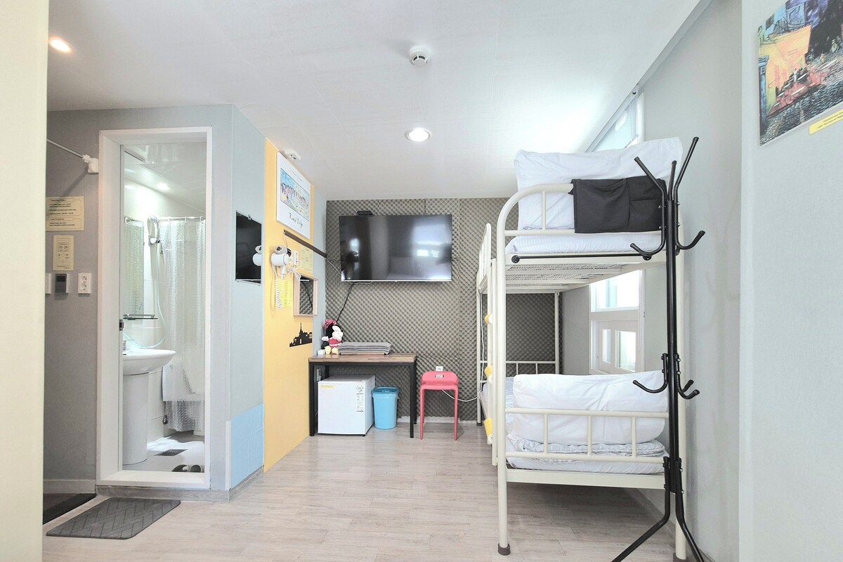 A room with a bunk bed, a desk with a computer, a small chair, and a TV on the wall.