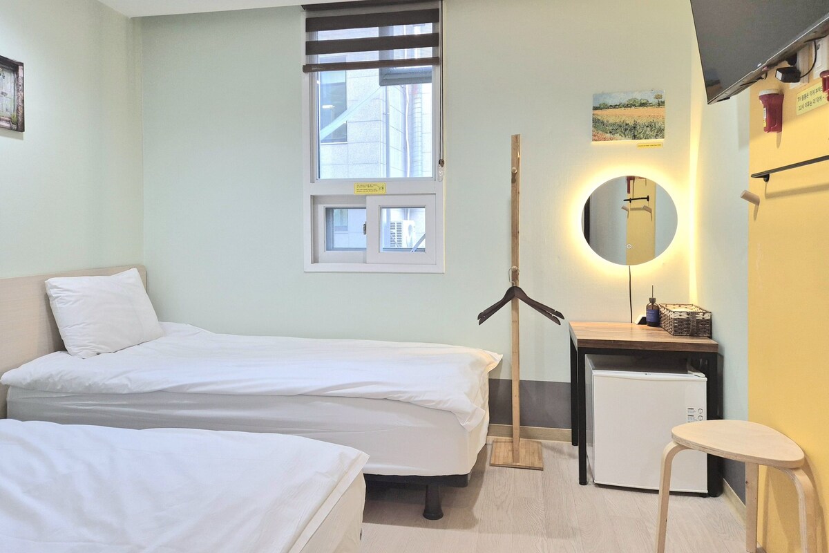 A hotel room with two beds, a small desk, a mirror, and a window with blinds.