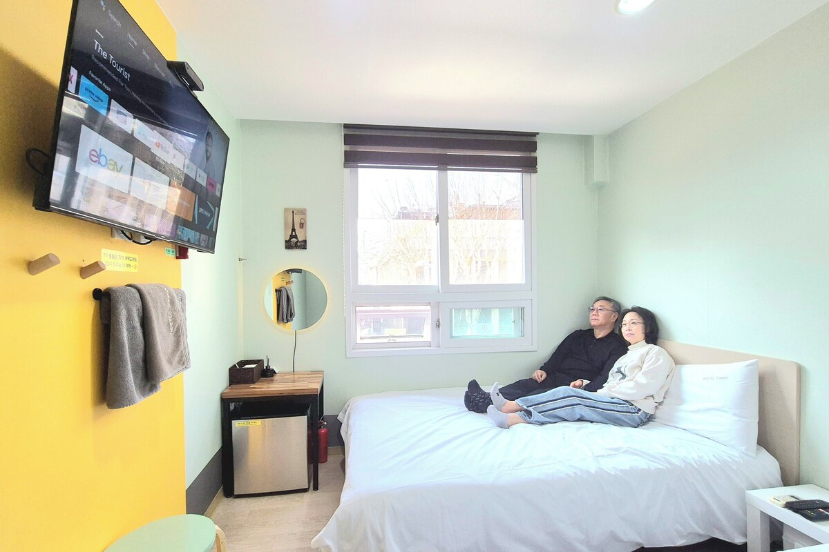 A bedroom with a bed, a TV mounted on the wall, a window, and a small table with a lamp.