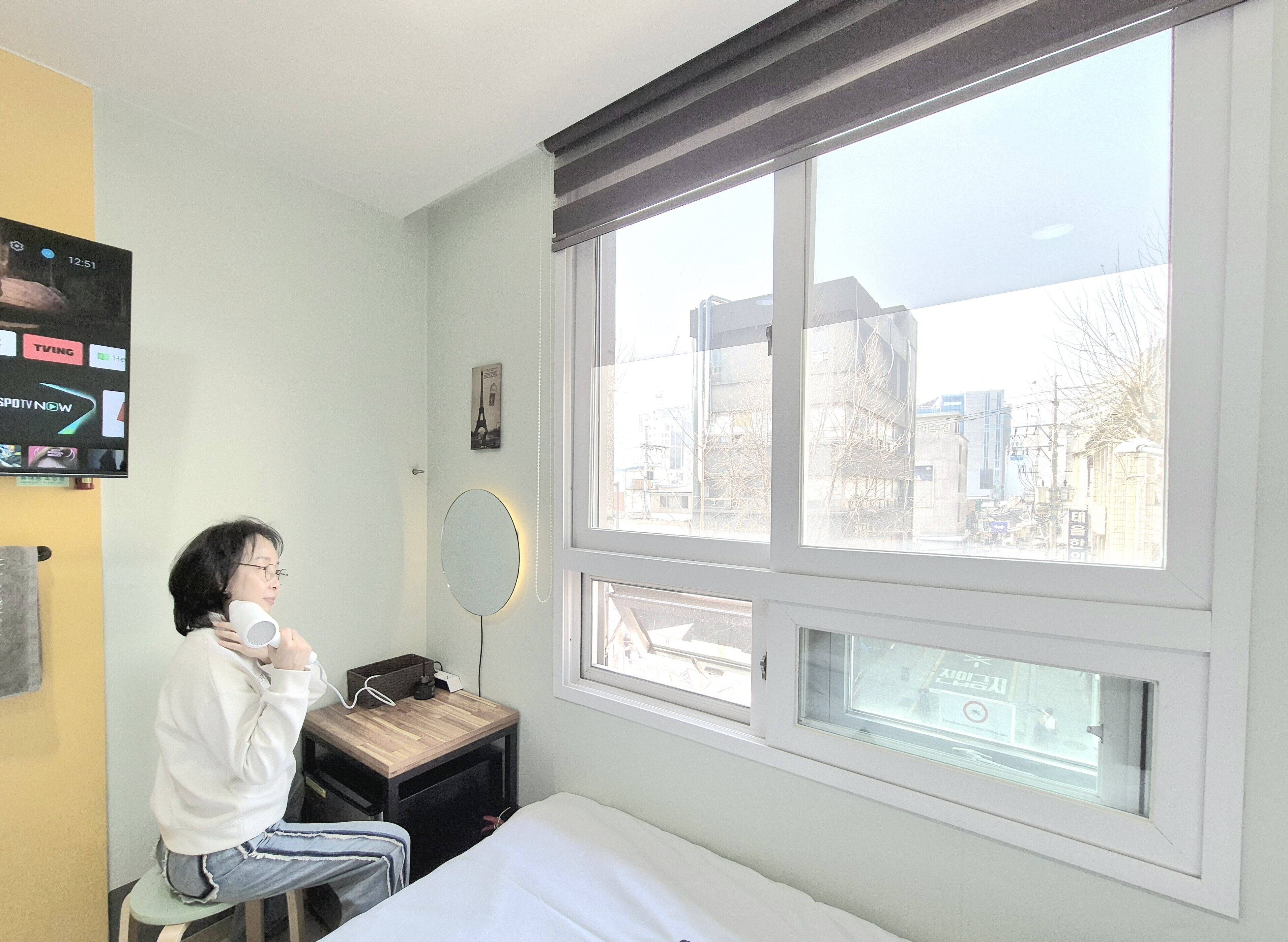 A hotel room with a bed, a bedside table with a lamp, a window with a view of buildings, and a TV on the wall.