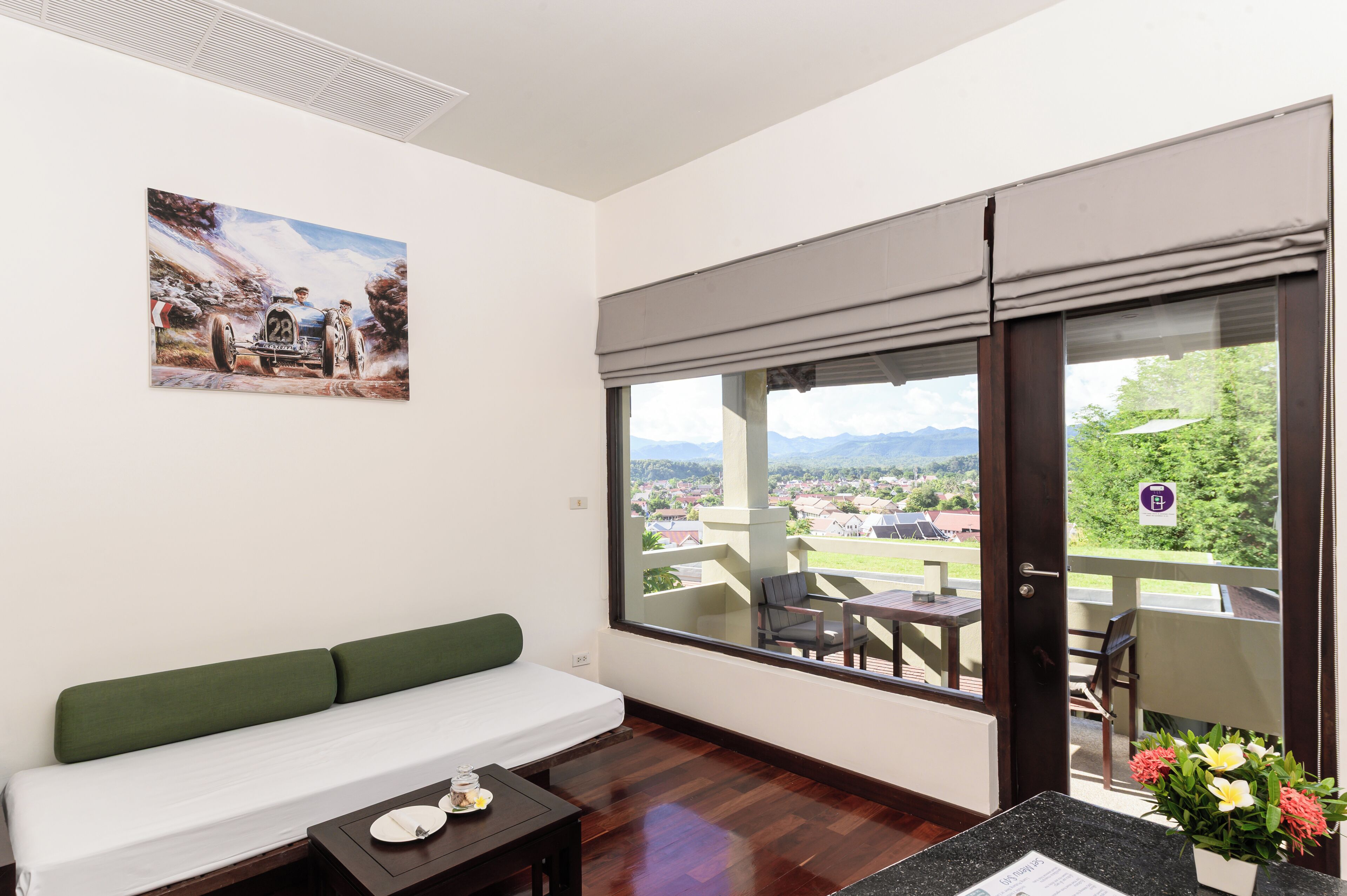 A room with a green sofa, a dining table with chairs, a painting on the wall, and a large window with a view of a town and mountains.