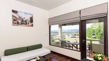 A room with a green sofa, a dining table with chairs, a painting on the wall, and a large window with a view of a town and mountains.