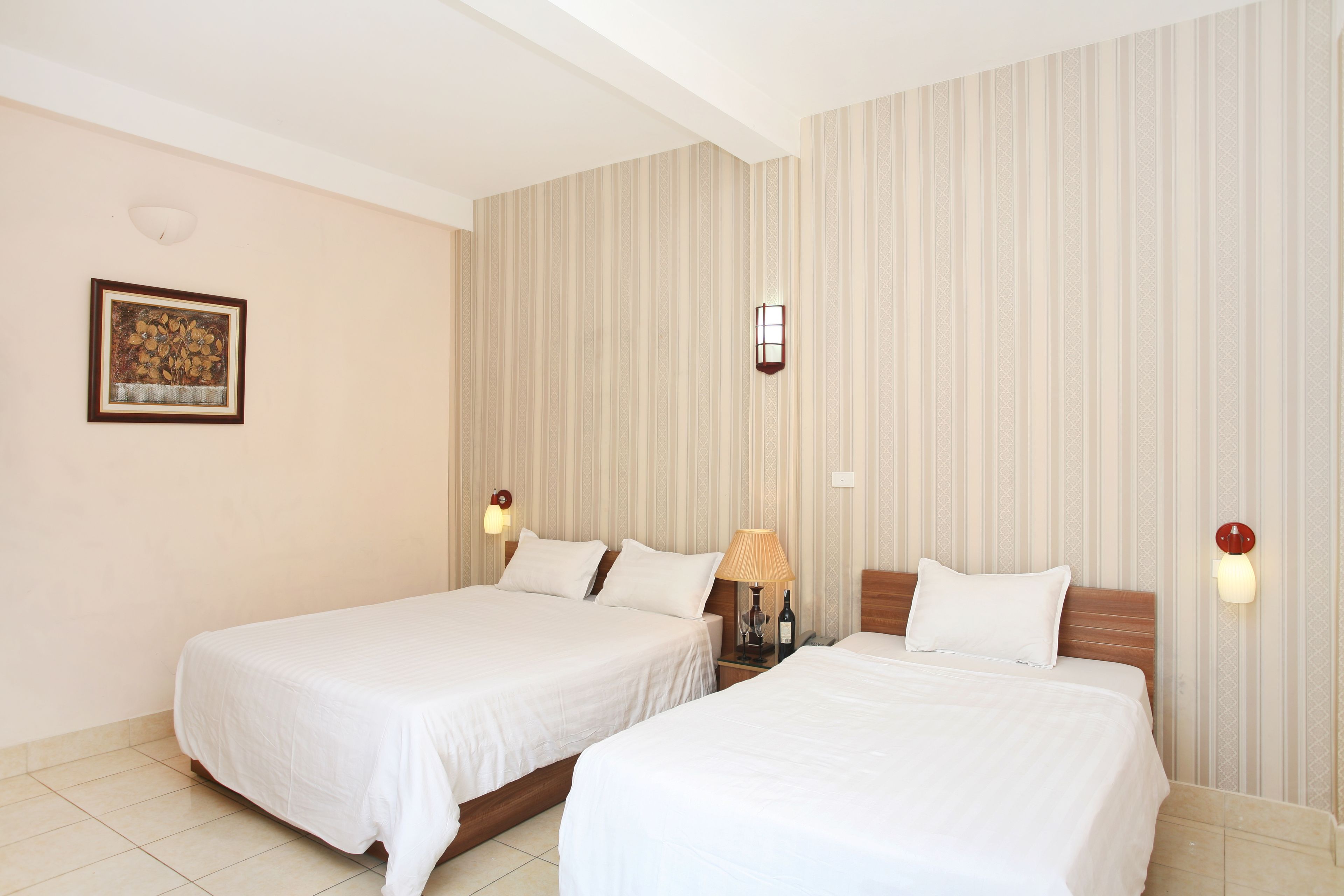 A hotel room with two single beds, a nightstand with a lamp, a framed picture on the wall, and a striped wallpaper design.