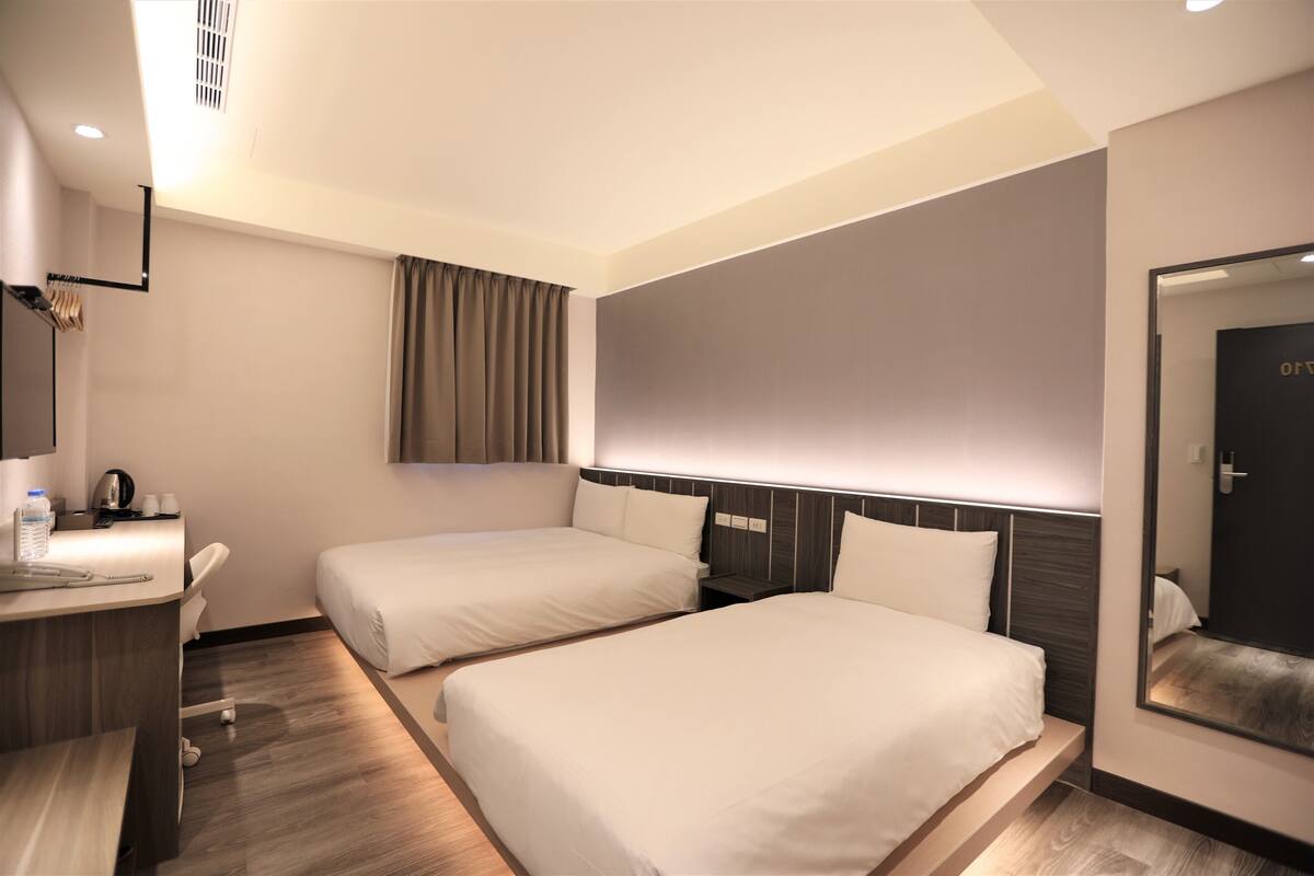 A hotel room with two beds, a desk, a mirror, and a window with curtains.