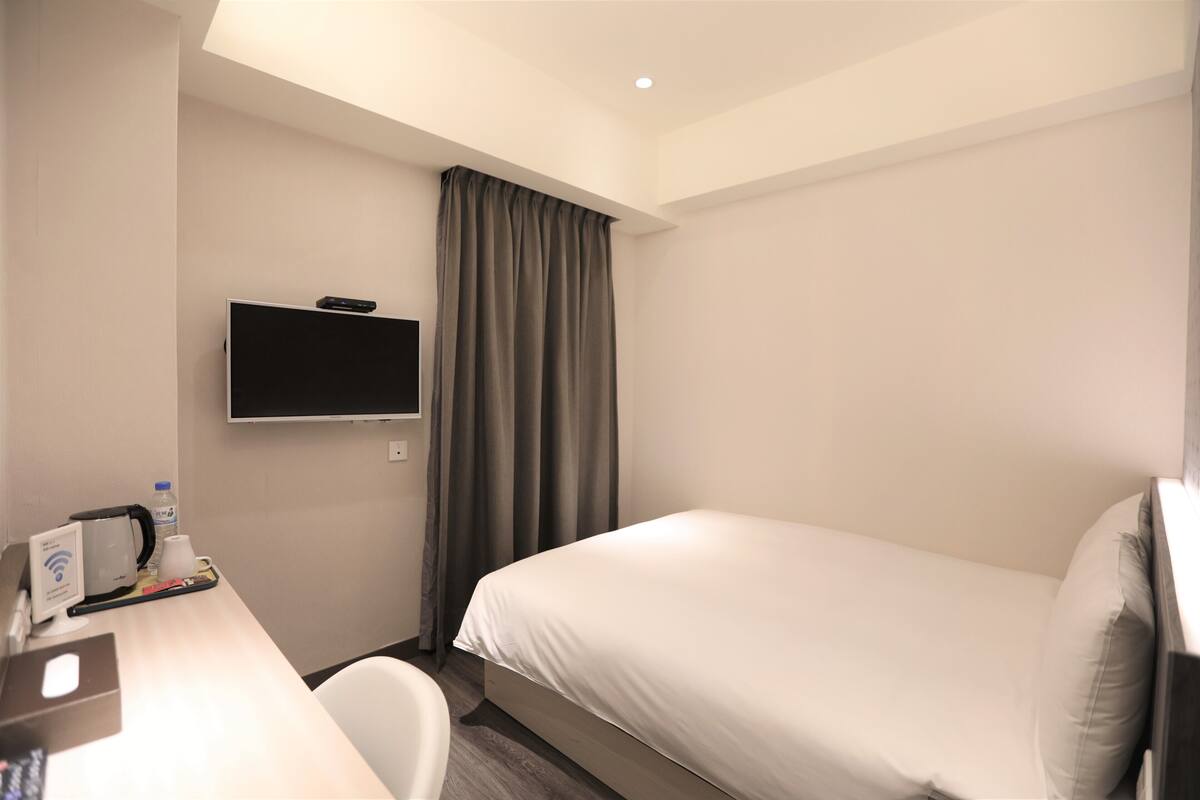 A hotel room with a bed, a desk with a coffee maker, a TV, and a window with curtains.