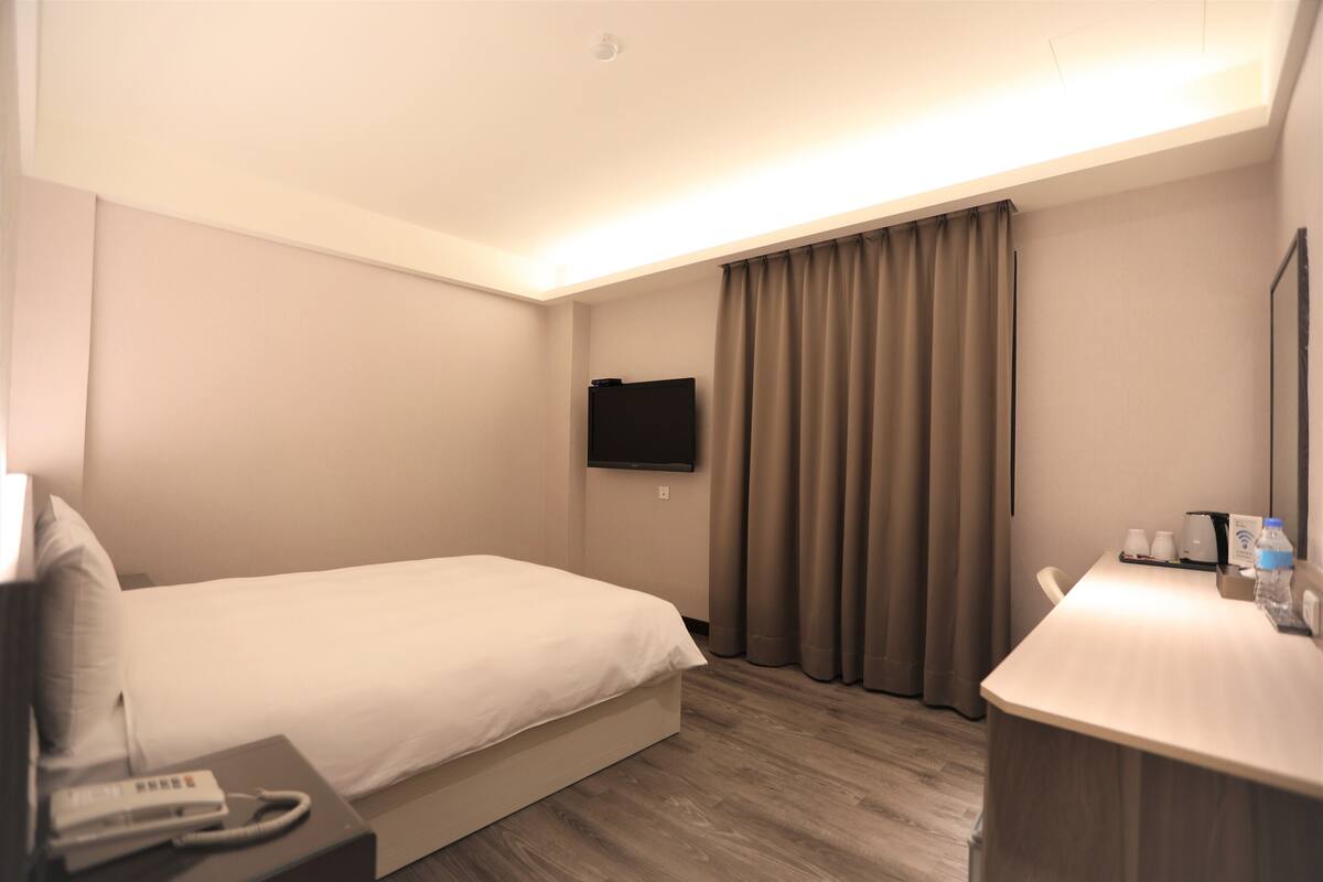 A hotel room with a bed, bedside table, a flat-screen TV mounted on the wall, a desk with a chair, and a window with curtains.