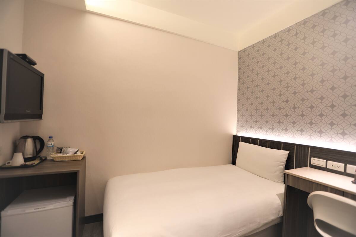 A hotel room with a bed, a desk with a kettle, a chair, and a television.