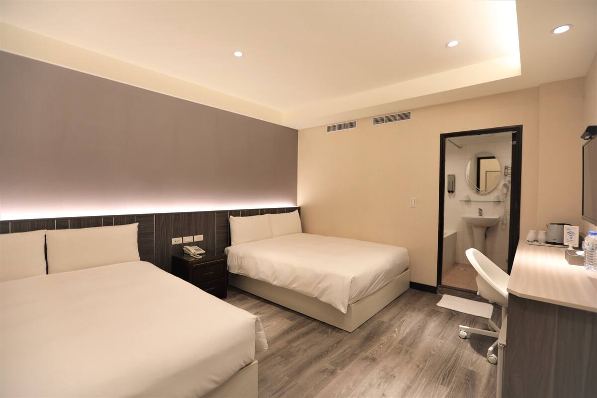 A hotel room with two beds, a desk, a chair, a mirror, and a bathroom visible through an open door.