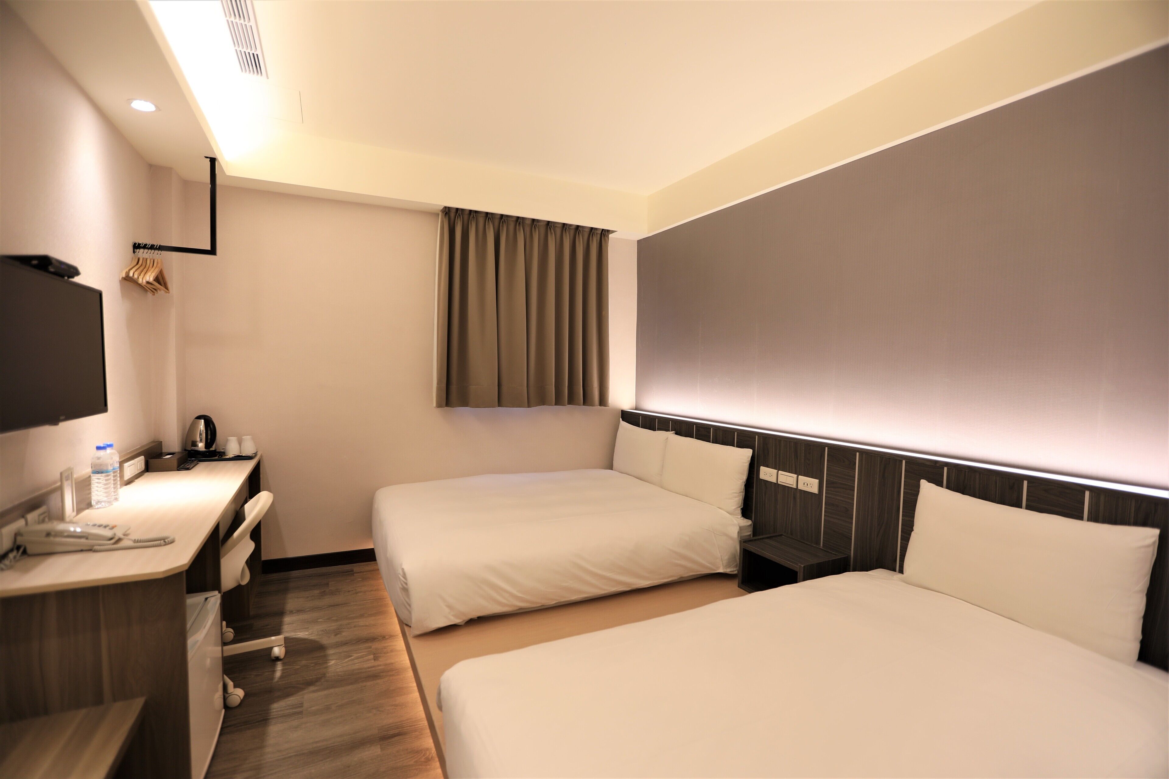 A hotel room with two beds, a desk with a telephone, a flat-screen TV, and a window with curtains.