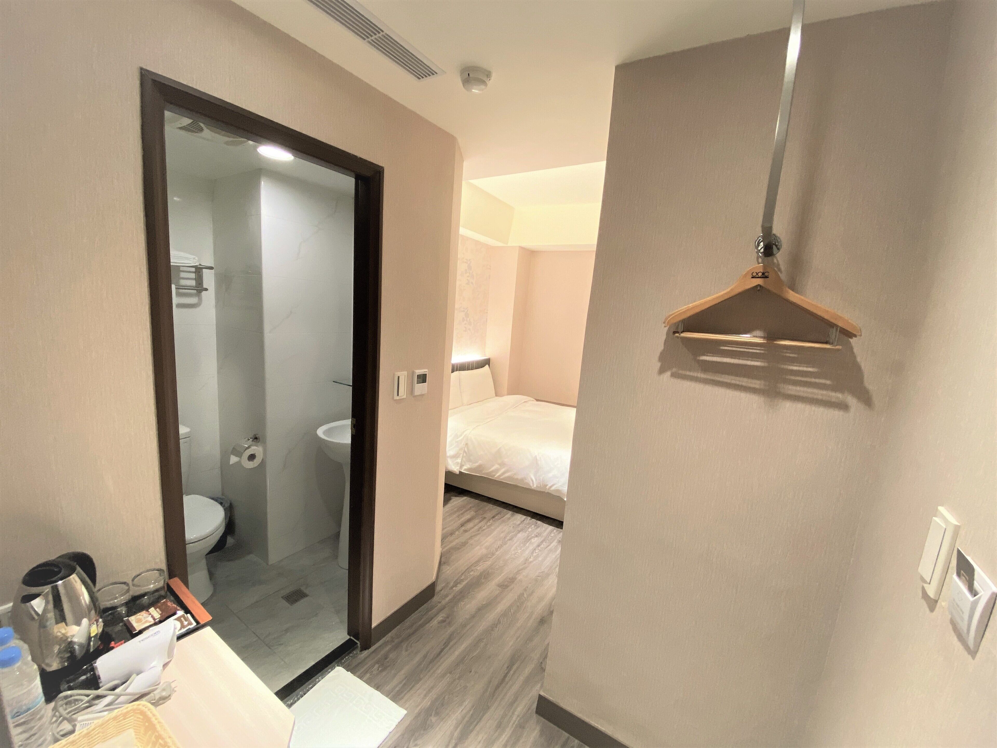 A hotel room with a bathroom, a bed, a bedside table with a lamp, and a hanging coat rack.