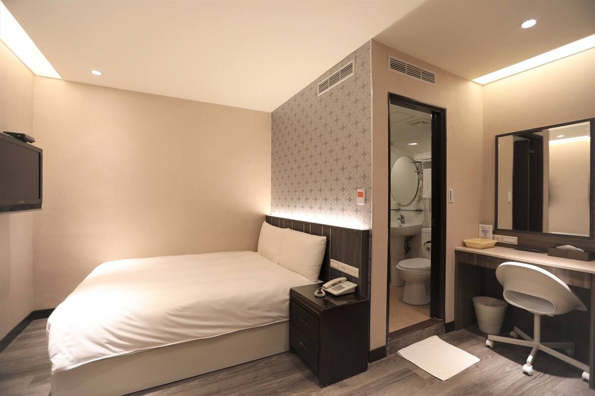 A hotel room with a bed, bedside table with a phone, a small trash bin, a desk with a chair, and a mirror.