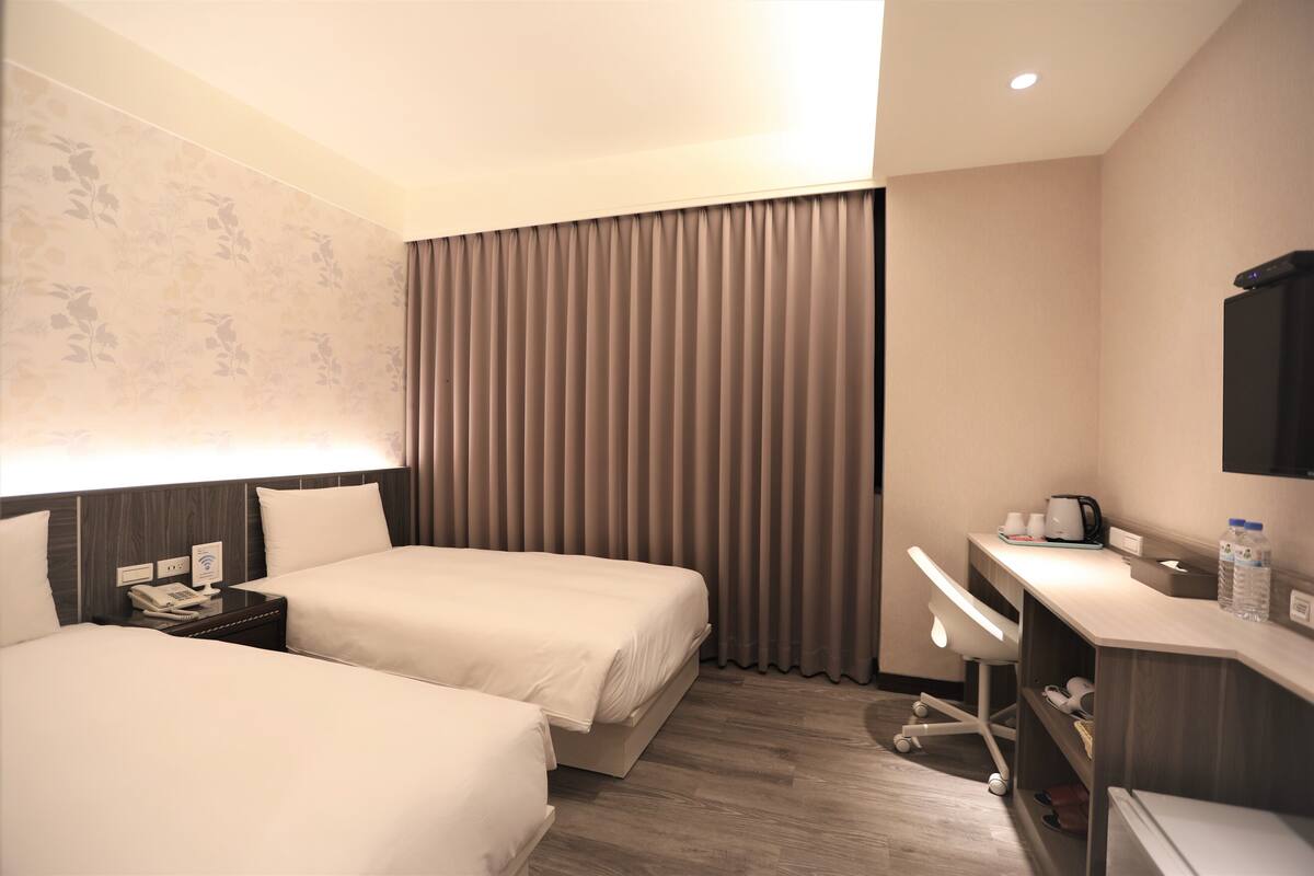 A hotel room with two beds, a desk, a chair, a telephone, and a flat-screen TV.