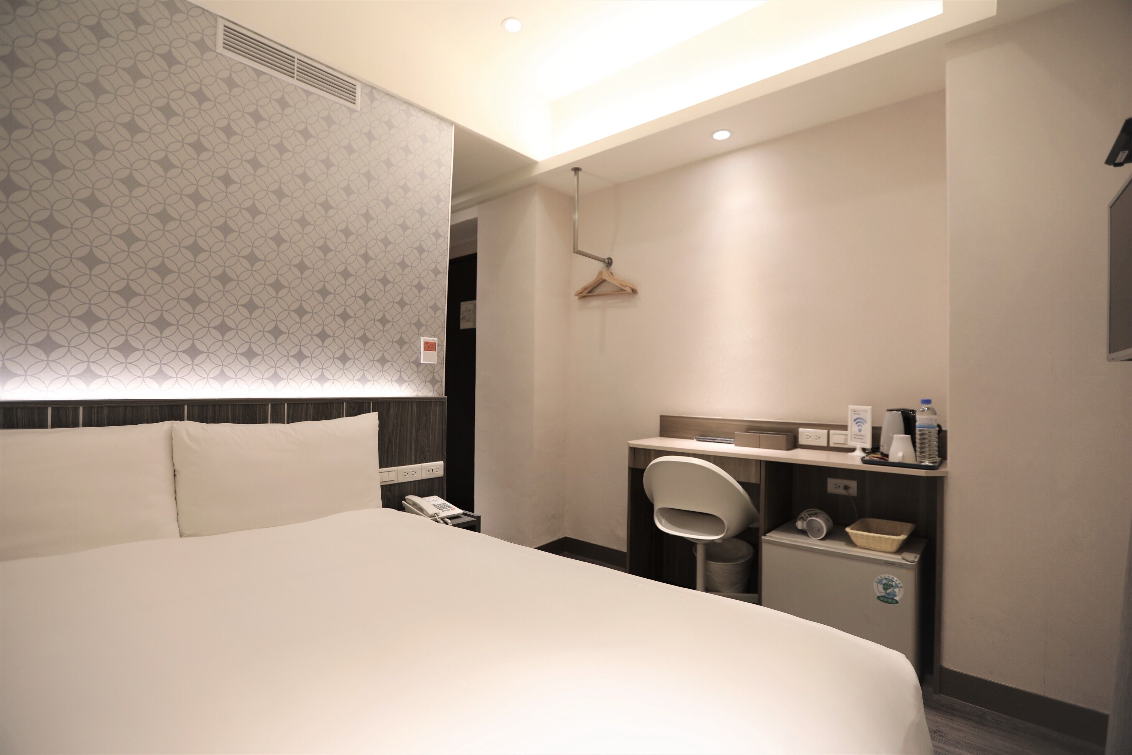 Hotellihuone, jossa on suuri sänky, työpöytä tuolilla, peili, pieni roskakori ja seinälle ripustettava naulakko.