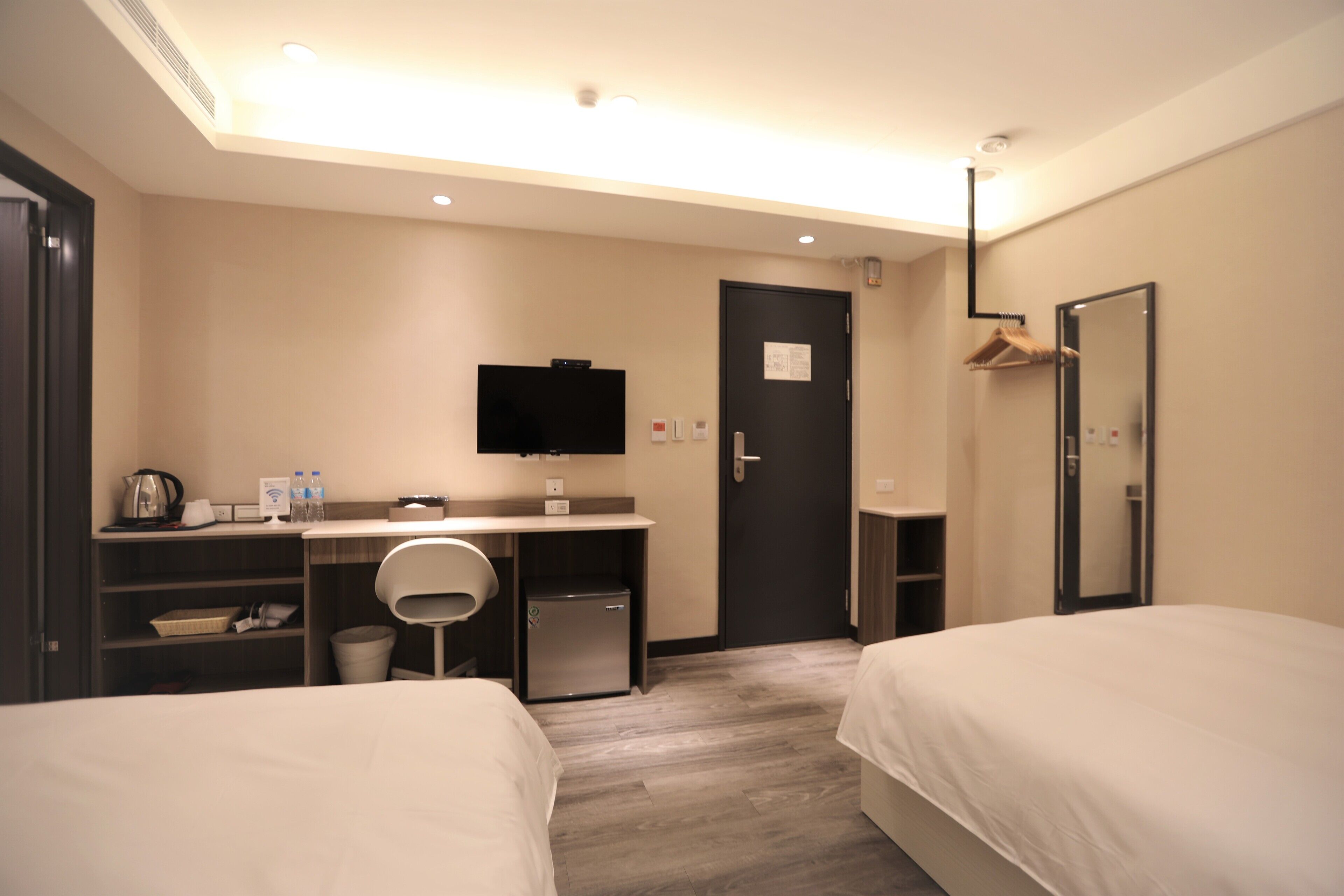 Quarto de hotel com duas camas, uma escrivaninha com computador e uma televisão de tela plana, uma pequena cozinha e um espelho.