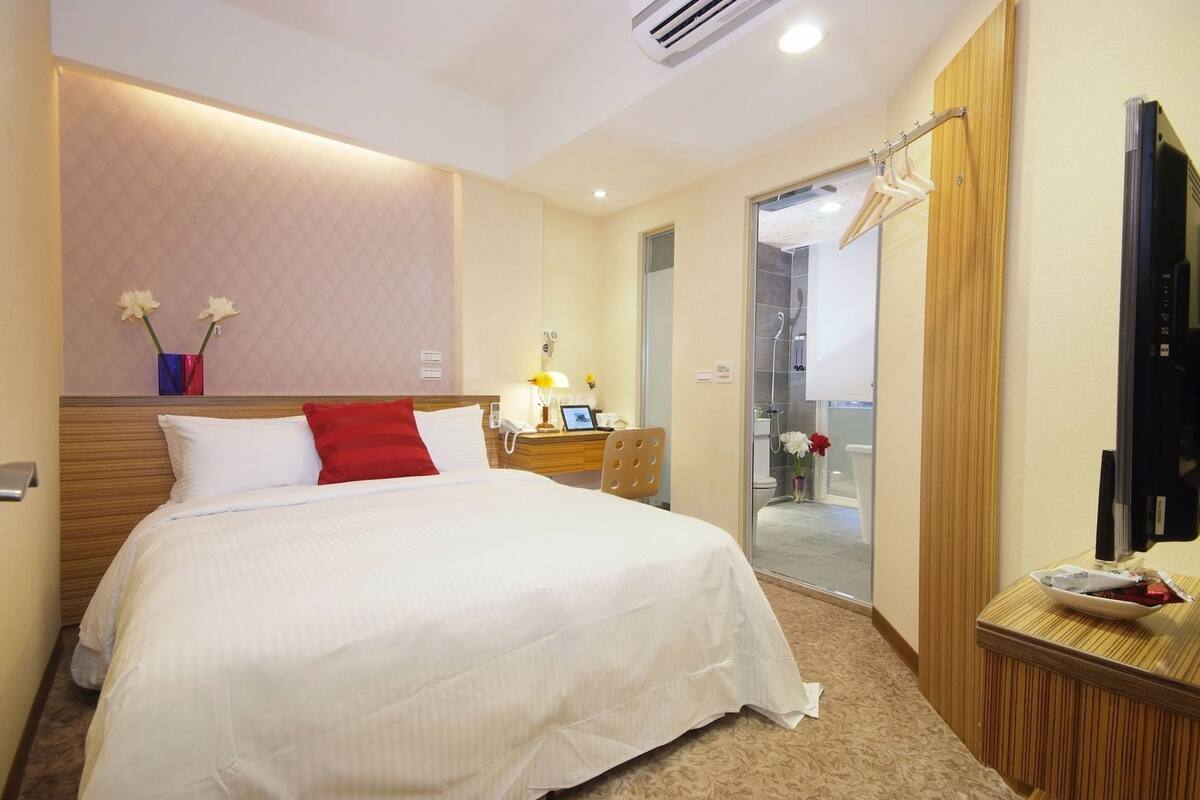 A hotel room with a bed, a nightstand, a mirror, a TV, and a shower area.