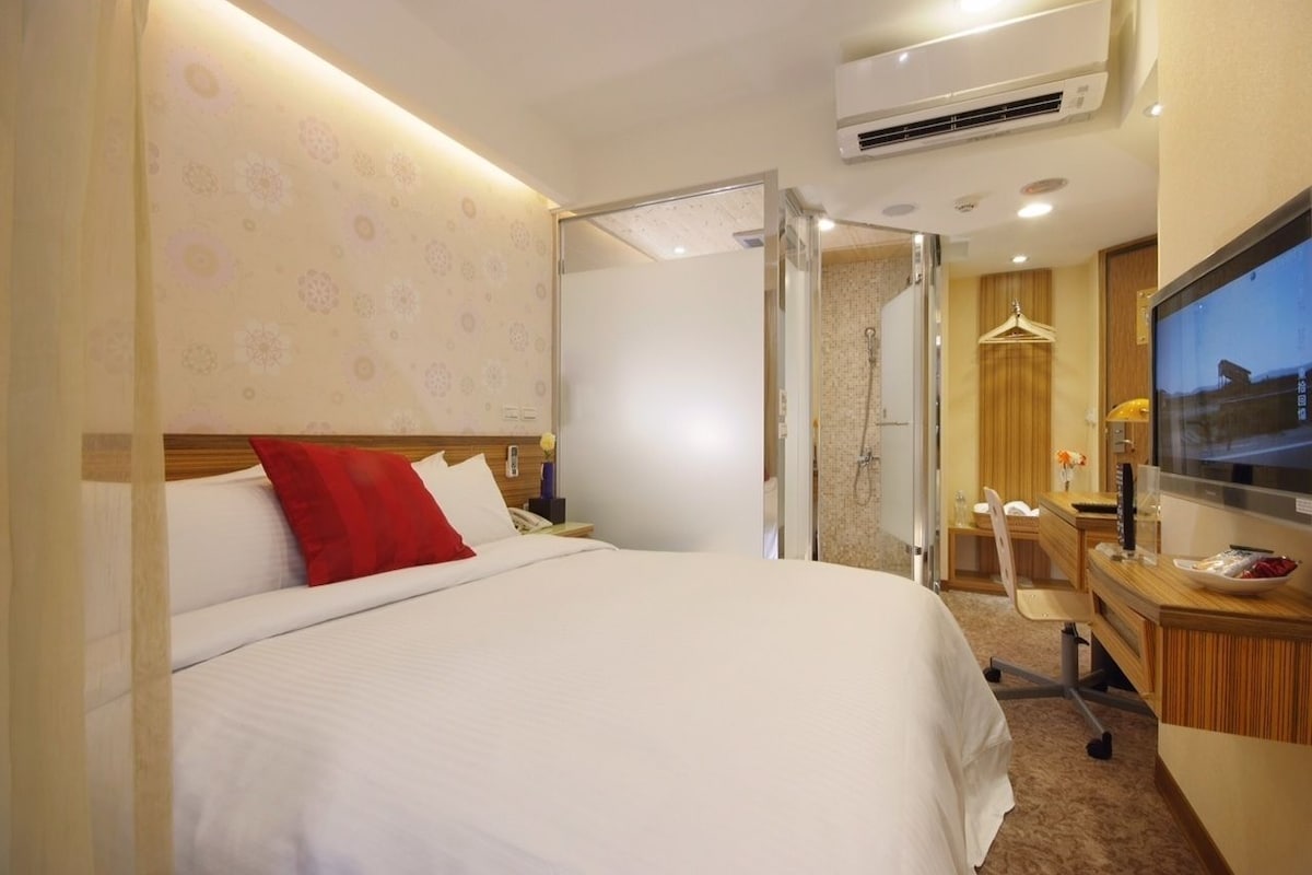 A hotel room with a bed, a desk, a TV, and an air conditioning unit.