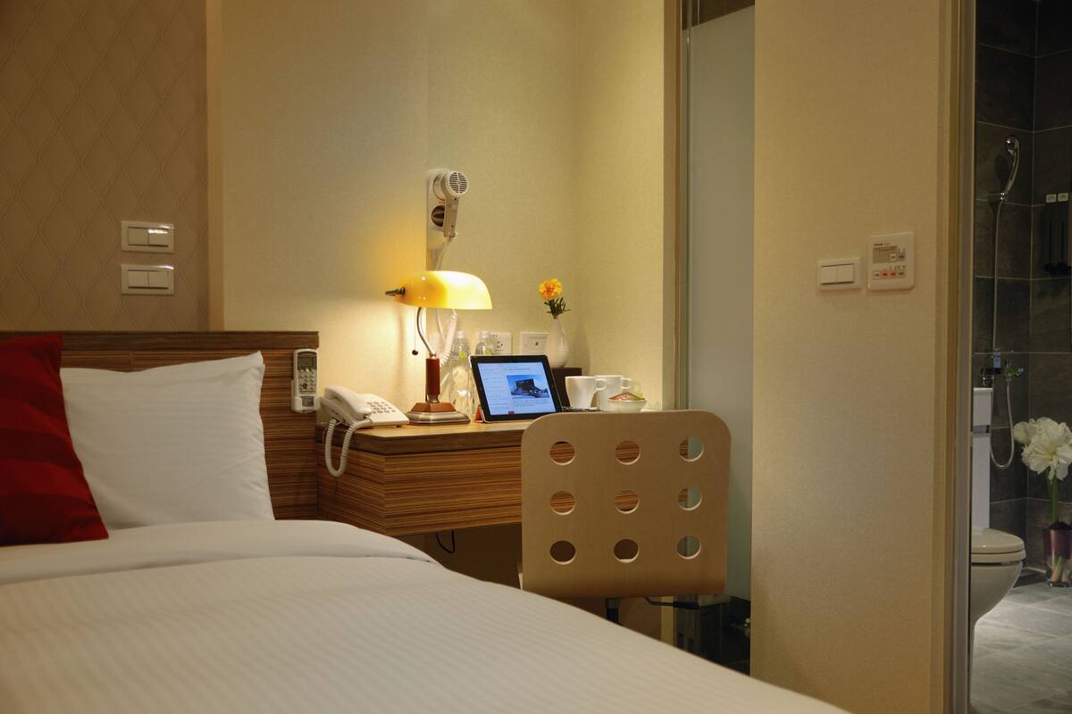 A hotel room with a bed, a desk with a lamp, a phone, a laptop, and a chair.