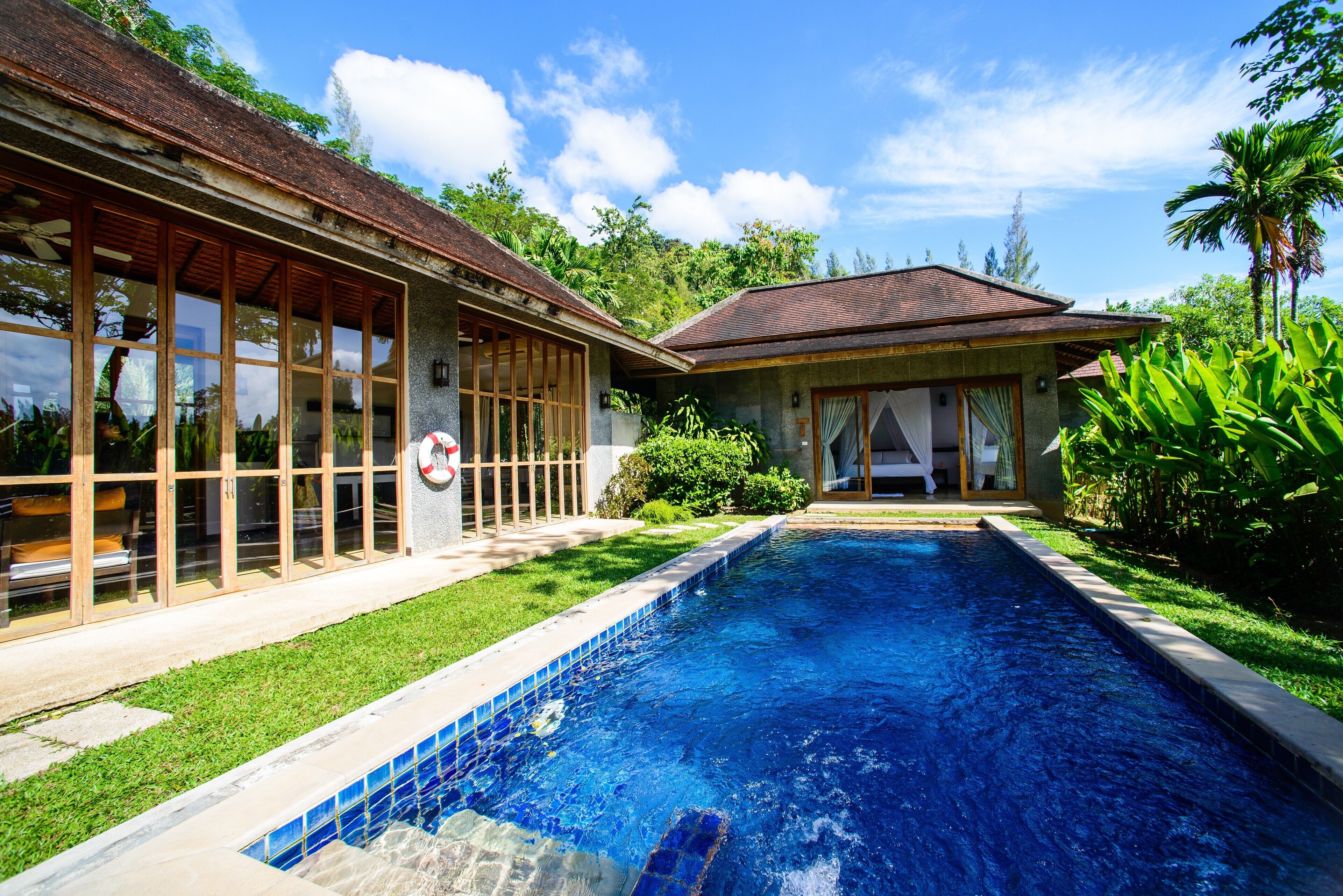 A swimming pool with a tiled edge, surrounded by a well-maintained lawn and a house with large windows and a tiled roof.