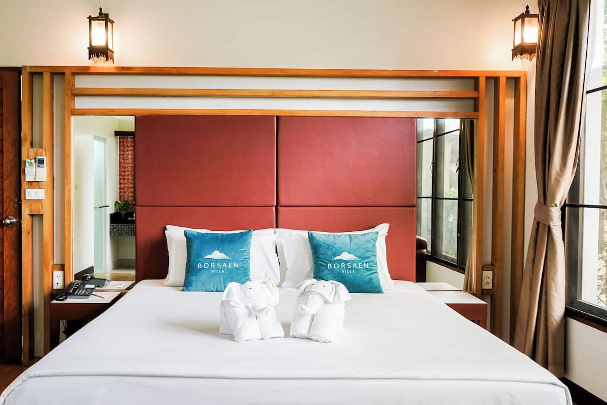 Ein Hotelzimmer mit einem großen Bett, zwei Kissen mit dem Borsaen Villa Logo, einem Nachttisch mit Telefon und Notizblock sowie einem Fenster mit Vorhängen.