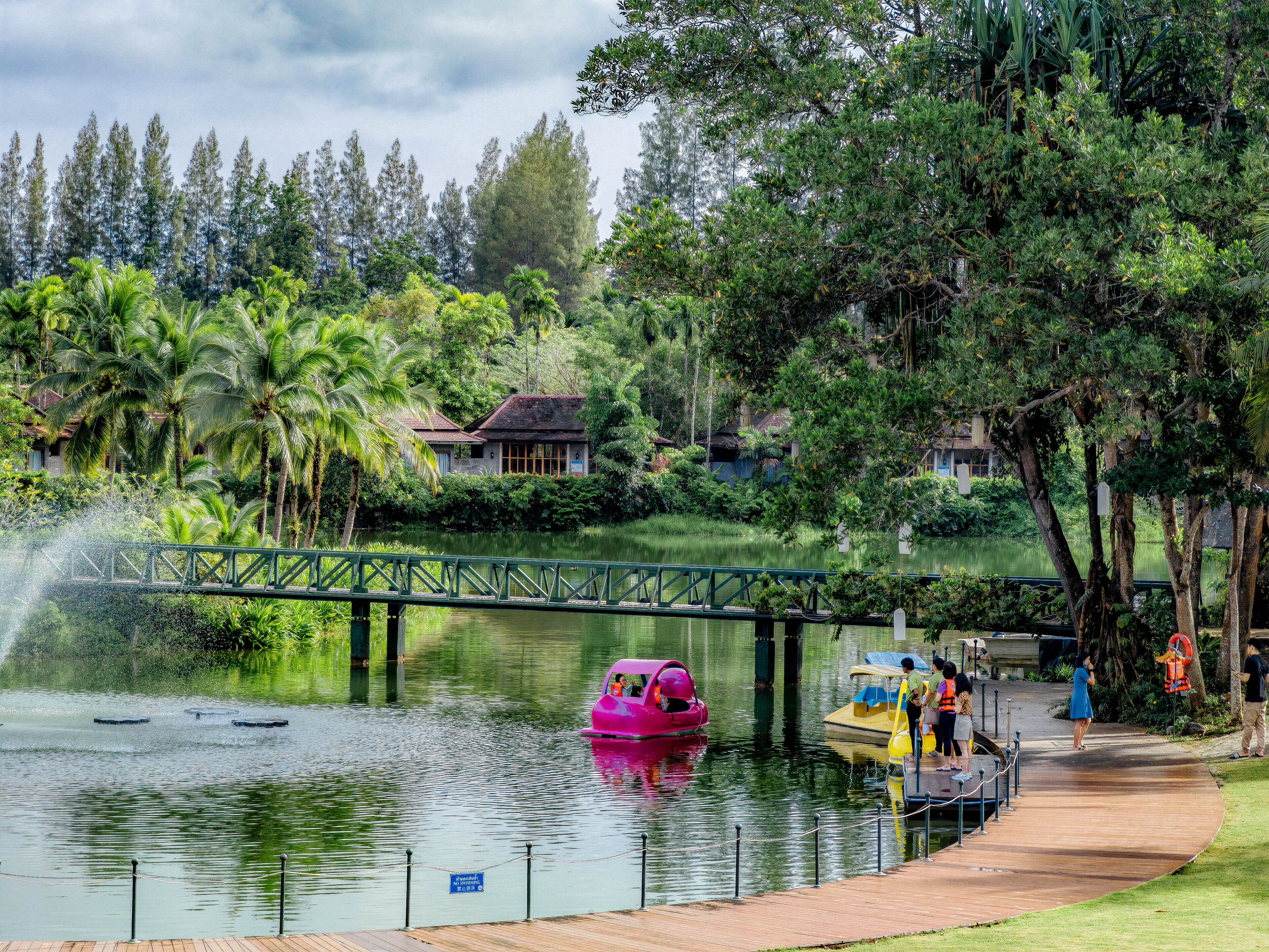 Cena tranquila à beira do lago com uma ponte, um barco rosa e um cais com pessoas.