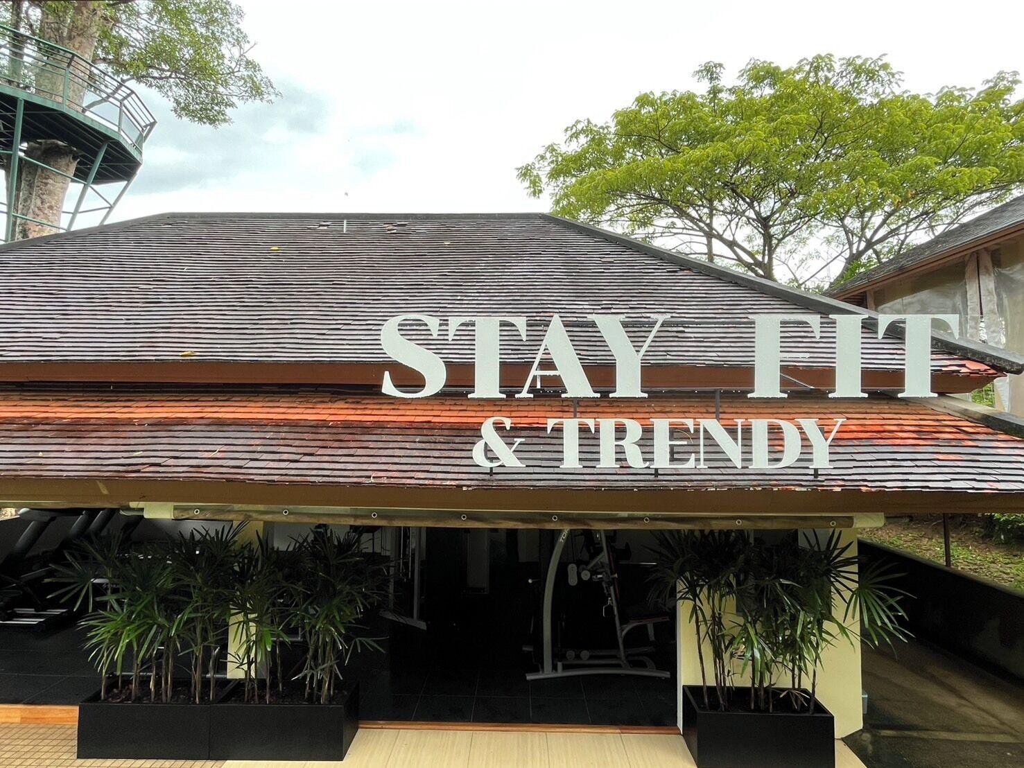 Letrero que dice 'STAY FIT & TRENDY', con techo inclinado y plantas en macetas al frente.