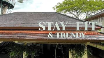 Letrero que dice 'STAY FIT & TRENDY', con techo inclinado y plantas en macetas al frente.