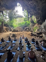 A group of people meditating in a cave with natural rock formations and greenery visible through an opening.