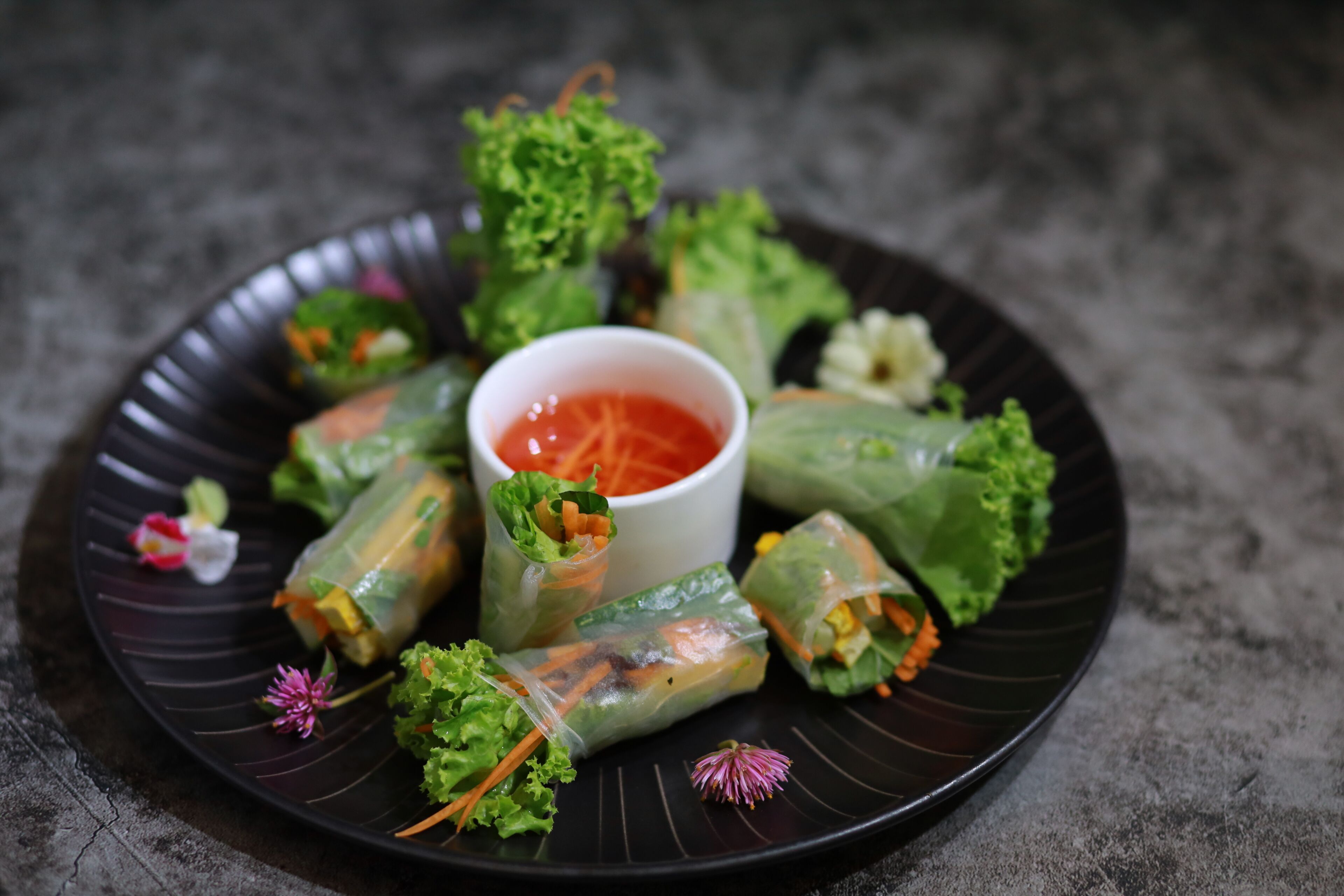 A plate of spring rolls with a dipping sauce in the center, surrounded by fresh greens and flowers.