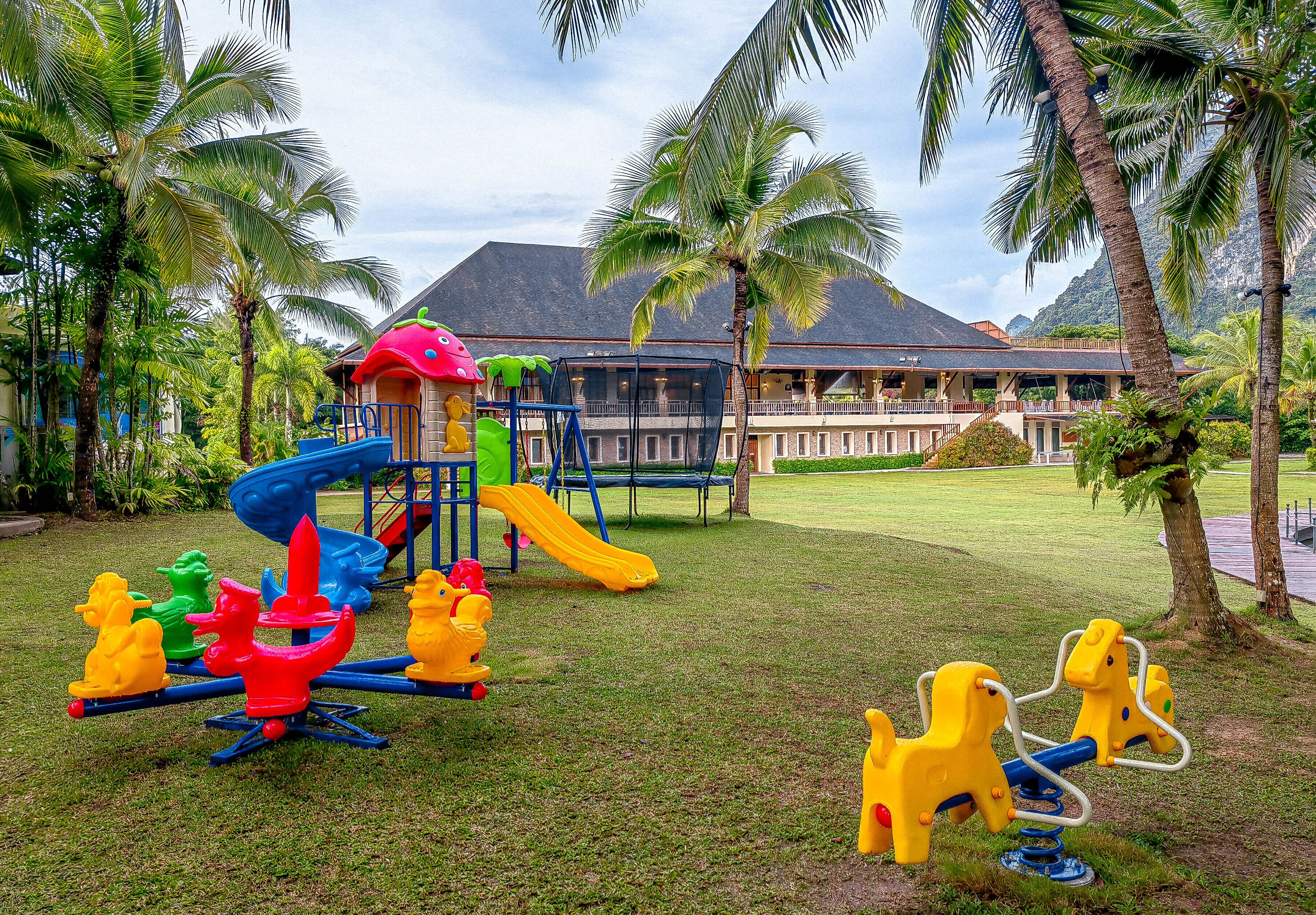 A playground with a slide and swings, surrounded by palm trees and a building with a red roof in the background.