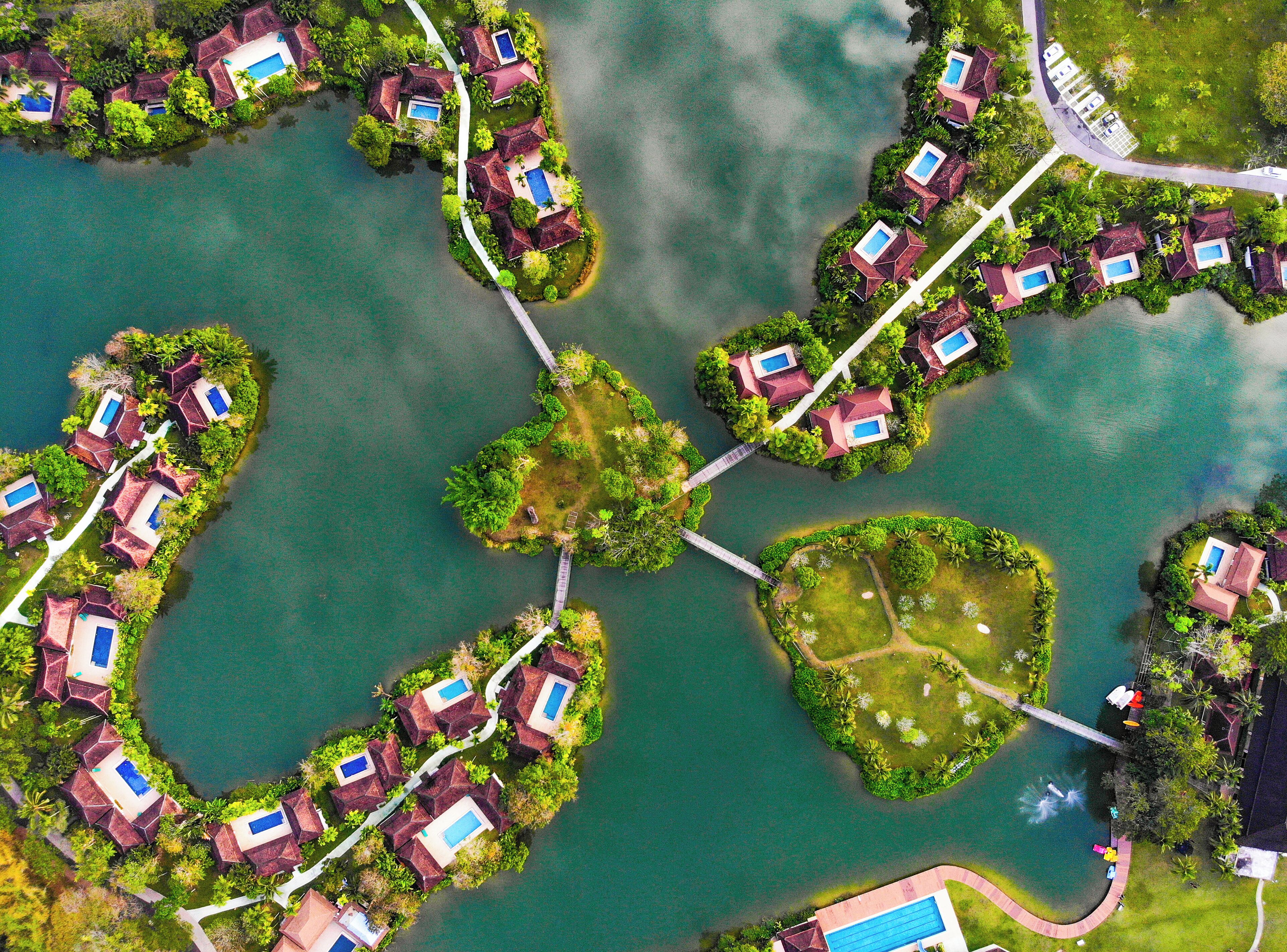 An aerial view of a resort with multiple islands connected by bridges, featuring villas with pools and lush greenery.
