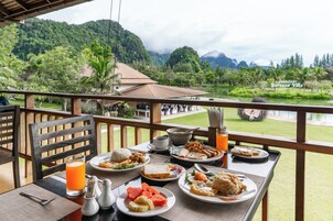 Una mesa de comedor con platos de comida, un vaso de jugo de naranja y vistas a un lago y montañas.
