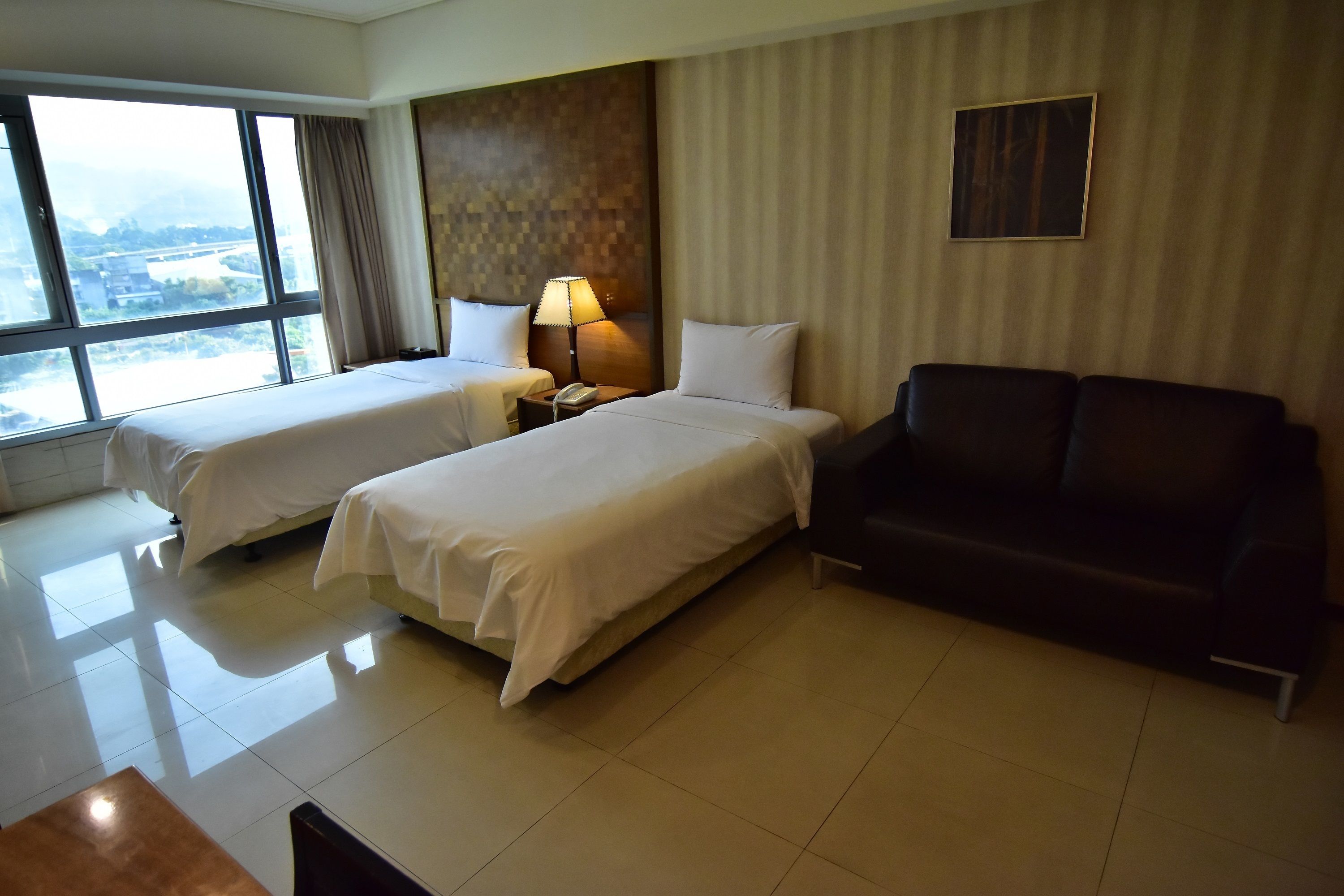 A hotel room with two beds, a sofa, a lamp, and a large window offering a city view.