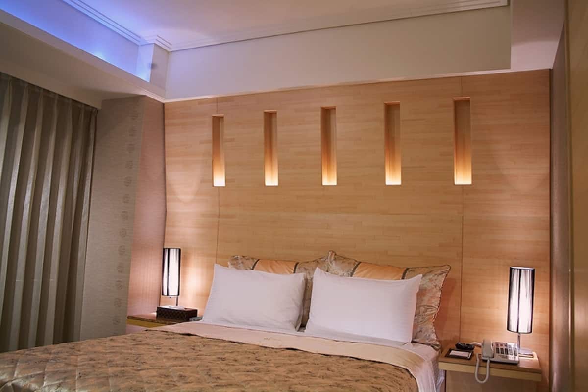 A hotel room with a large bed, a wooden headboard with built-in lighting, a bedside table with a lamp, and a window with curtains.