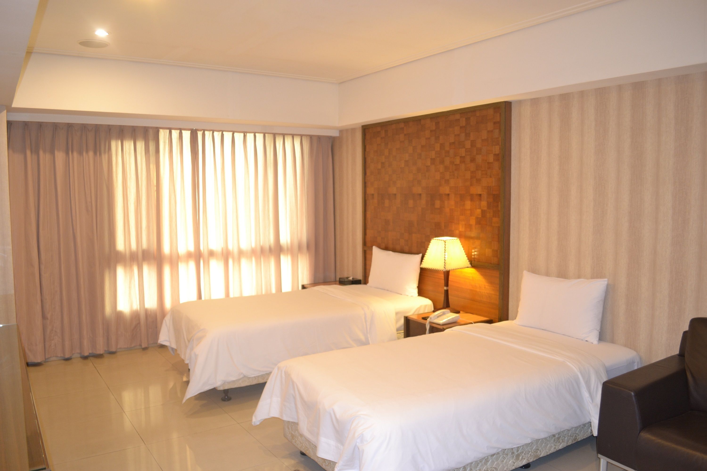 A hotel room with two beds, a nightstand with a lamp, a chair, and a large window with curtains.