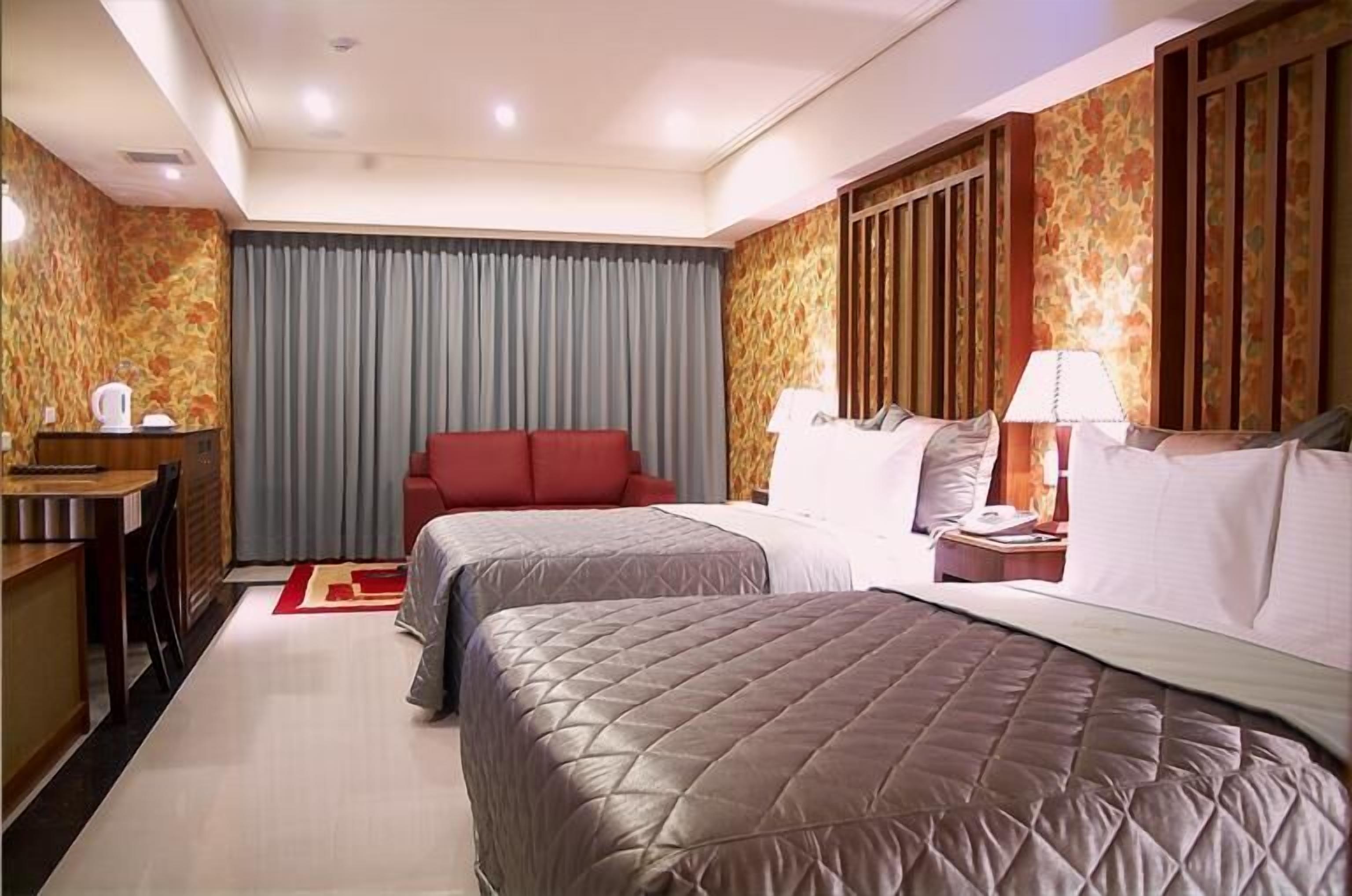 Ein Hotelzimmer mit zwei Betten, einem Schreibtisch, einem Stuhl, einem roten Sofa und einem Vorhang.