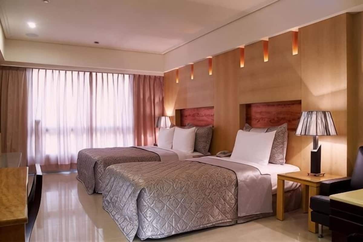 A hotel room with two beds, a desk, a chair, a lamp, and a large window with curtains.