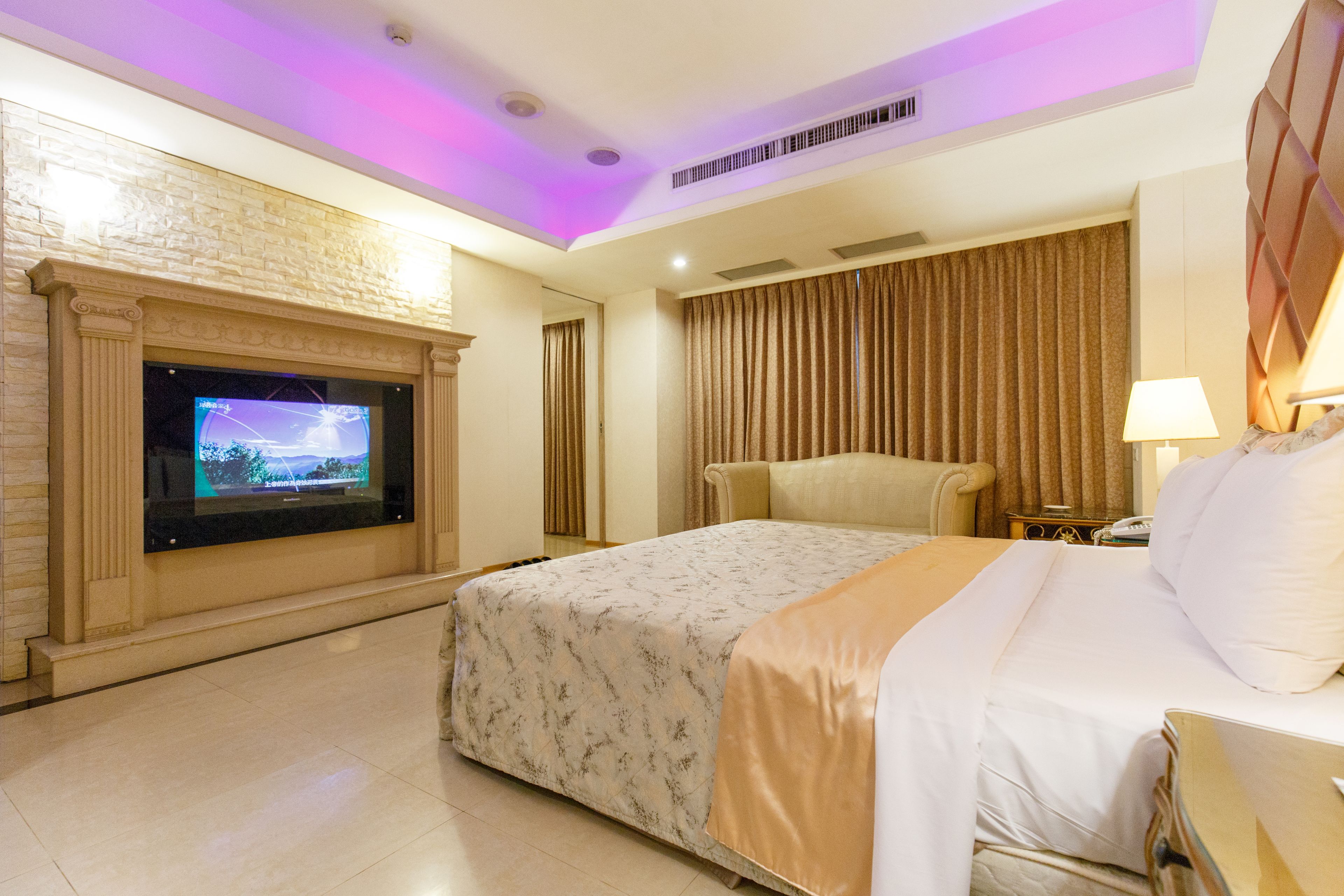 A hotel room with a large bed, a television mounted on a brick wall, a sofa, and a lamp on a bedside table.