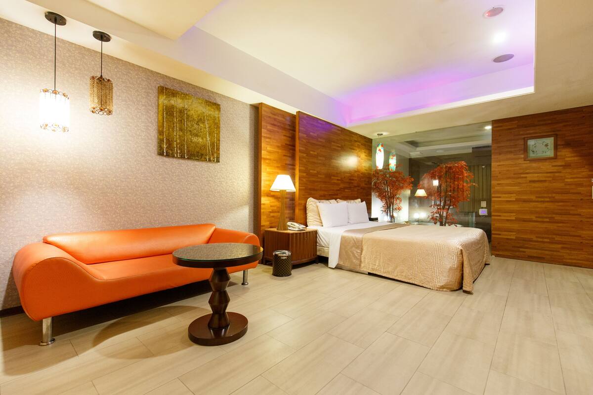 A hotel room with a modern design, featuring a wooden accent wall, a bed with a white comforter, an orange sofa, and a small round table.