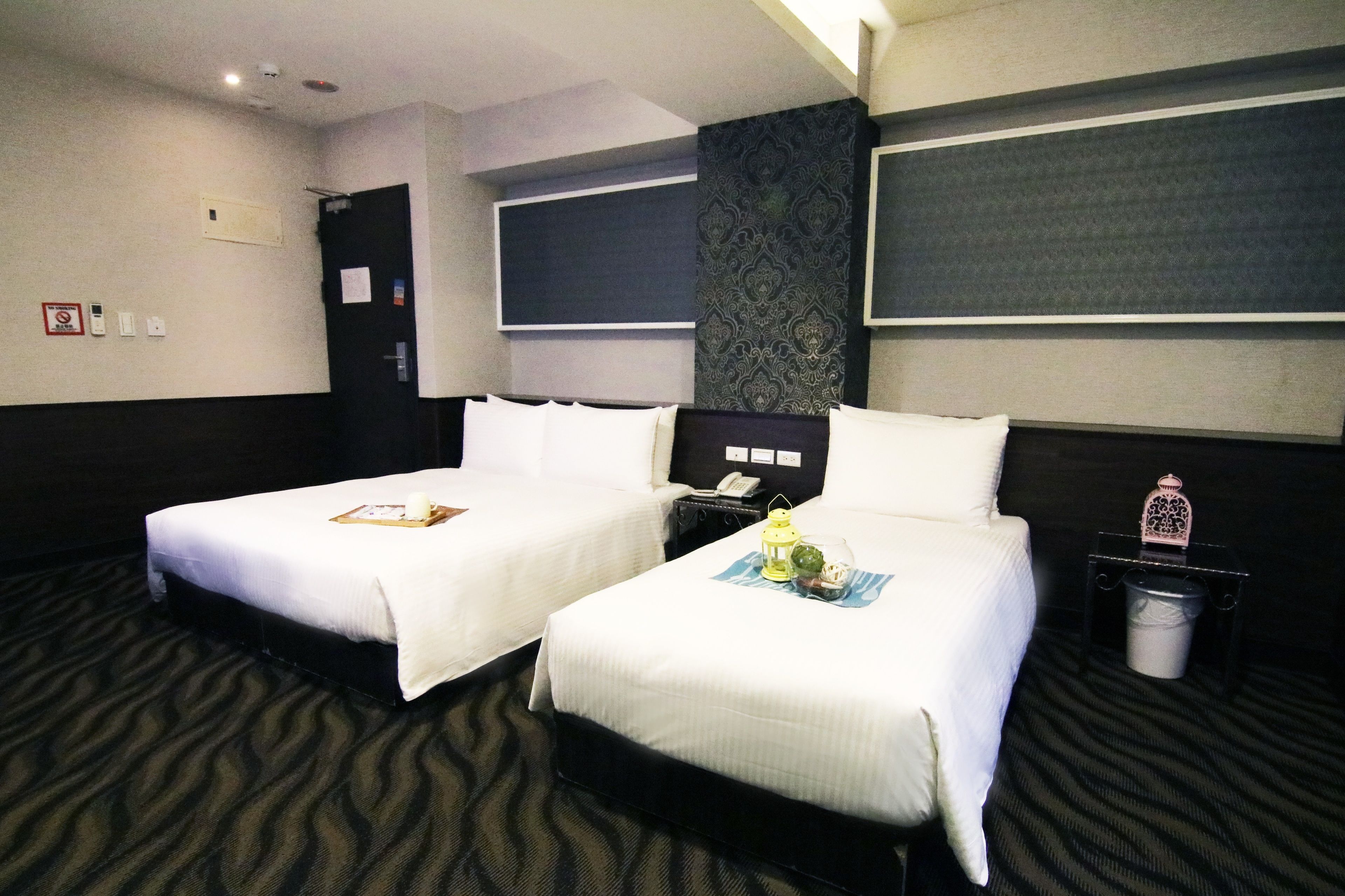 A hotel room with two beds, a dark patterned wall, a small trash bin, and a nightstand with a phone and a lantern.