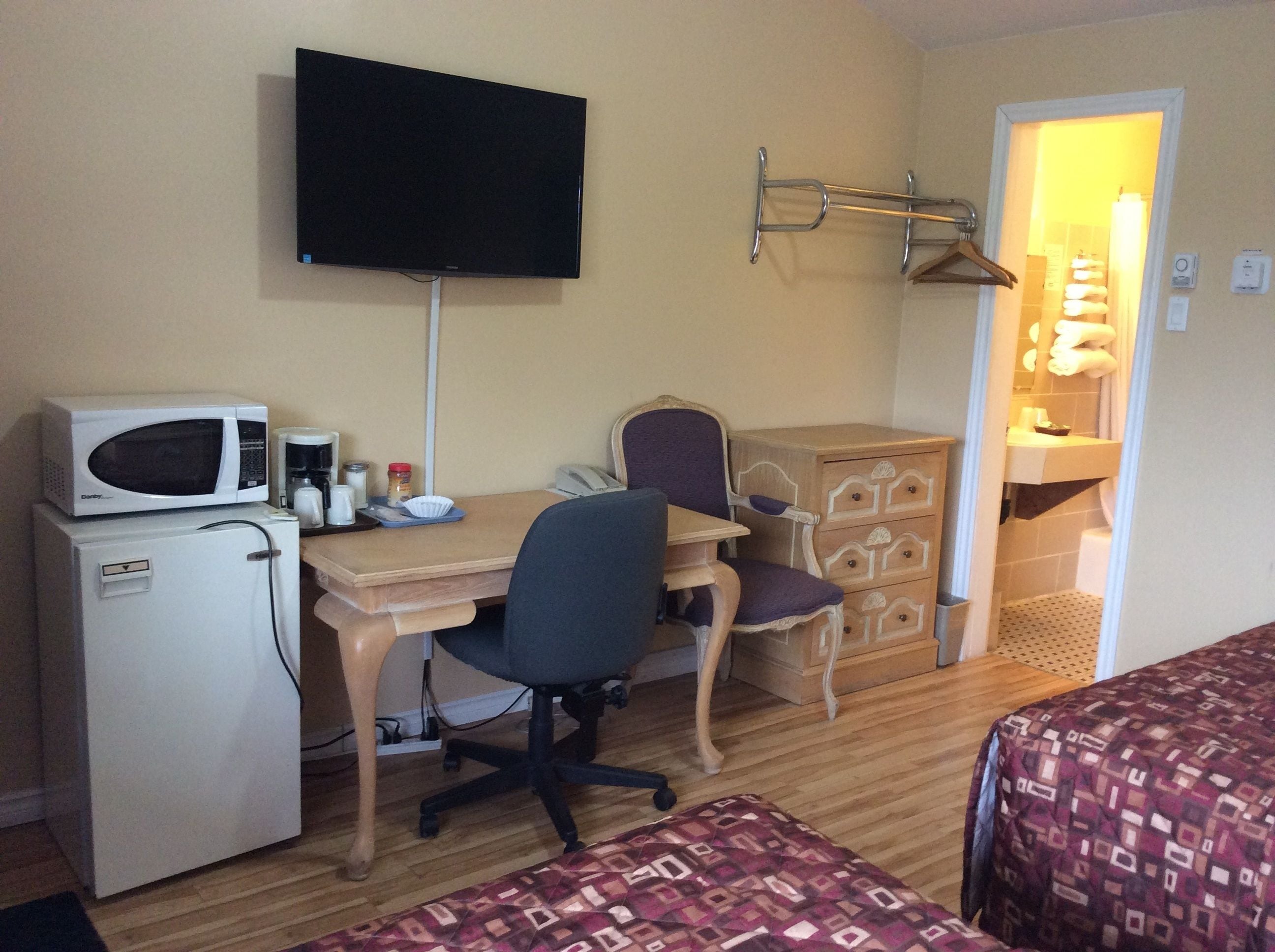 Quarto de hotel compacto com uma cama de solteiro, uma escrivaninha com cadeira, micro-ondas, mini-geladeira, televisão fixada na parede e cômoda de madeira.