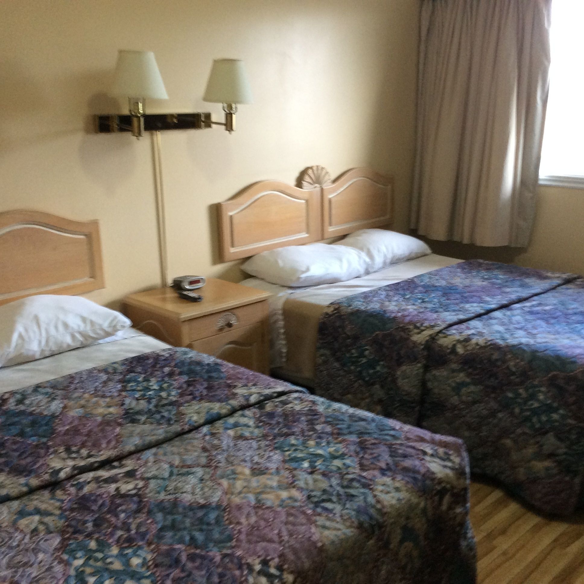 A hotel room with two beds, each with a floral bedspread, a wooden headboard, a nightstand with a lamp, and a window with curtains.