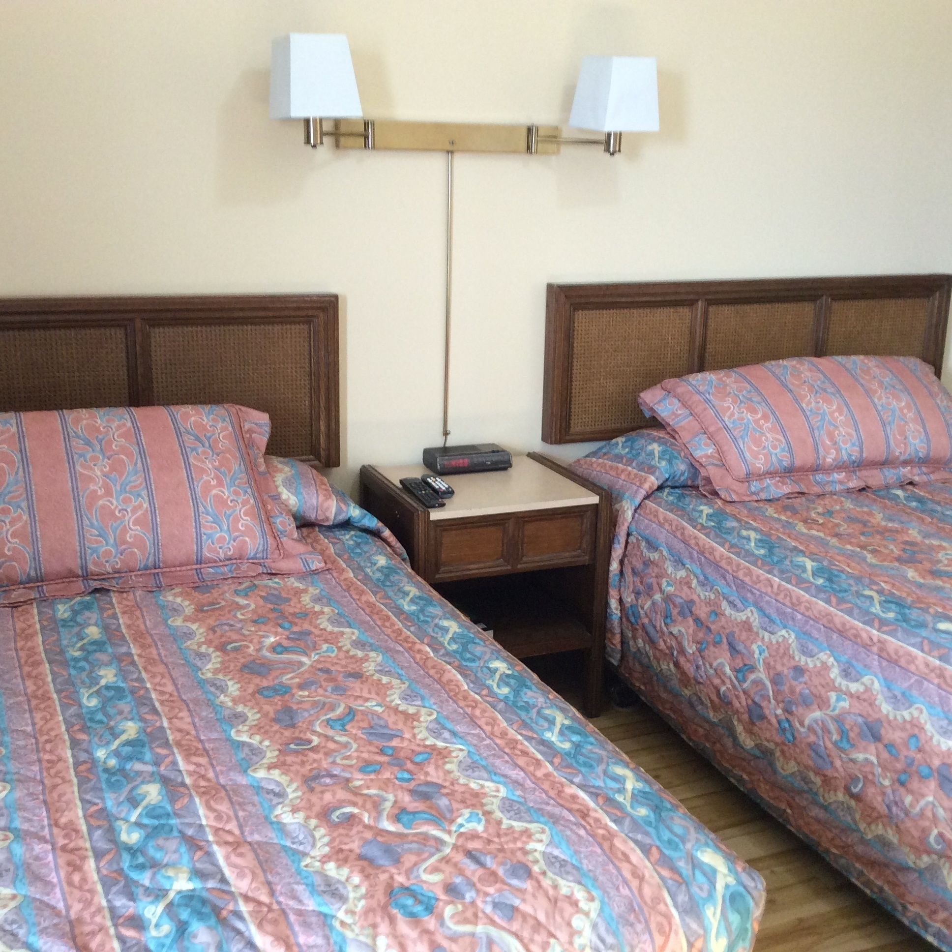 A hotel room with two beds, each with patterned bedding, a nightstand with a remote control, and wall-mounted lamps.