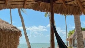 Escena de playa con una terraza de madera, una hamaca y una estructura de techo de paja.