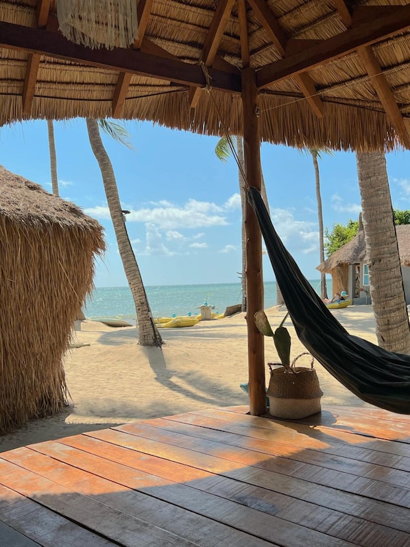 Escena de playa con una terraza de madera, una hamaca y una estructura de techo de paja.