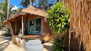 Una cabaña con techo de paja, puerta de vidrio, un pequeño espacio de estar al aire libre y una escalera que conduce a la entrada.