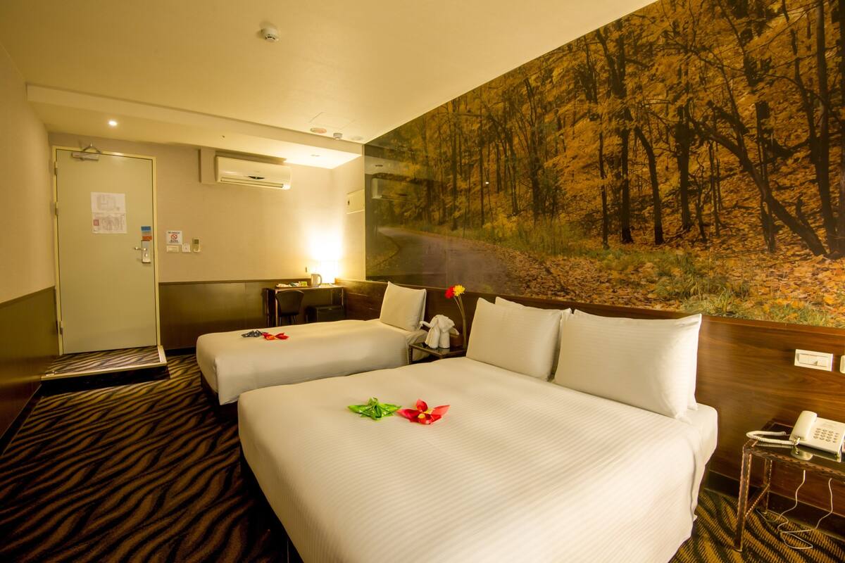 A hotel room with two beds, a painting of a forest on the wall, a desk with a lamp, and a telephone on a side table.
