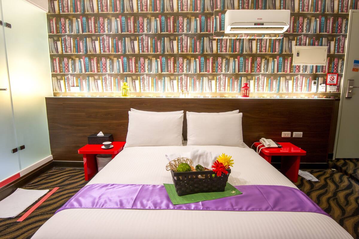 A hotel room with a large bed, two nightstands, a bookshelf, and a red side table.