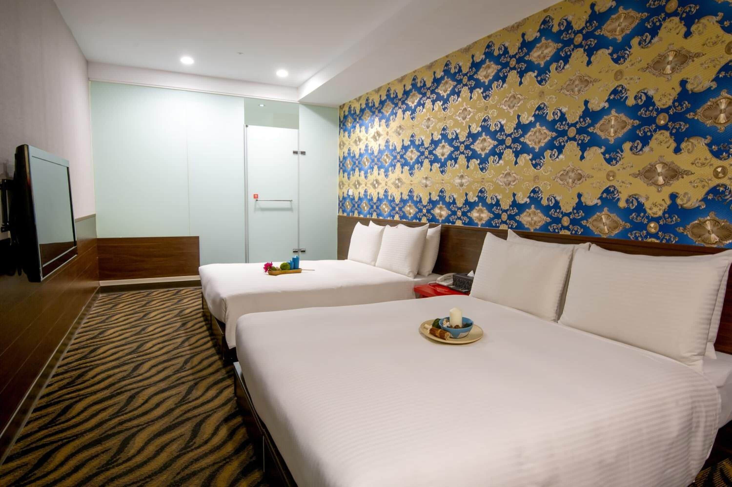 A hotel room with two beds, a flat-screen TV, a glass shower enclosure, and a patterned wallpaper.