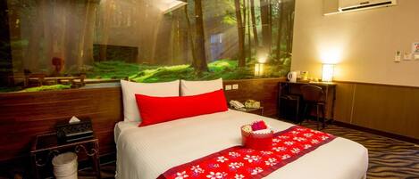 A hotel room with a large forest mural on the wall, a neatly made bed with a red pillow, a small desk with a lamp, and a trash bin.